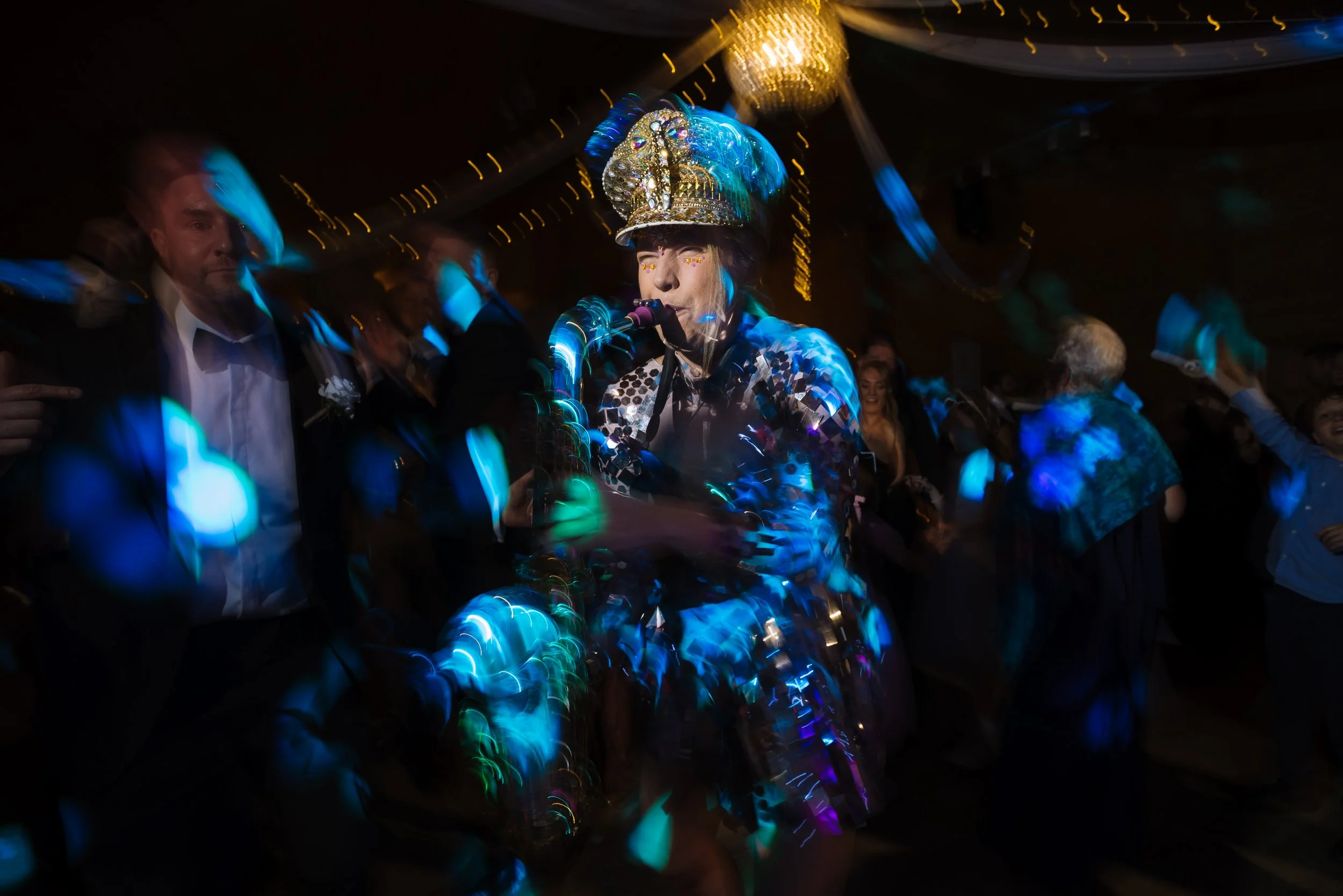 A woman dressed in a colorful costume with a shiny, feathered hat, playing saxaphone at a party with dancing guests, colorful lights, and decorations.