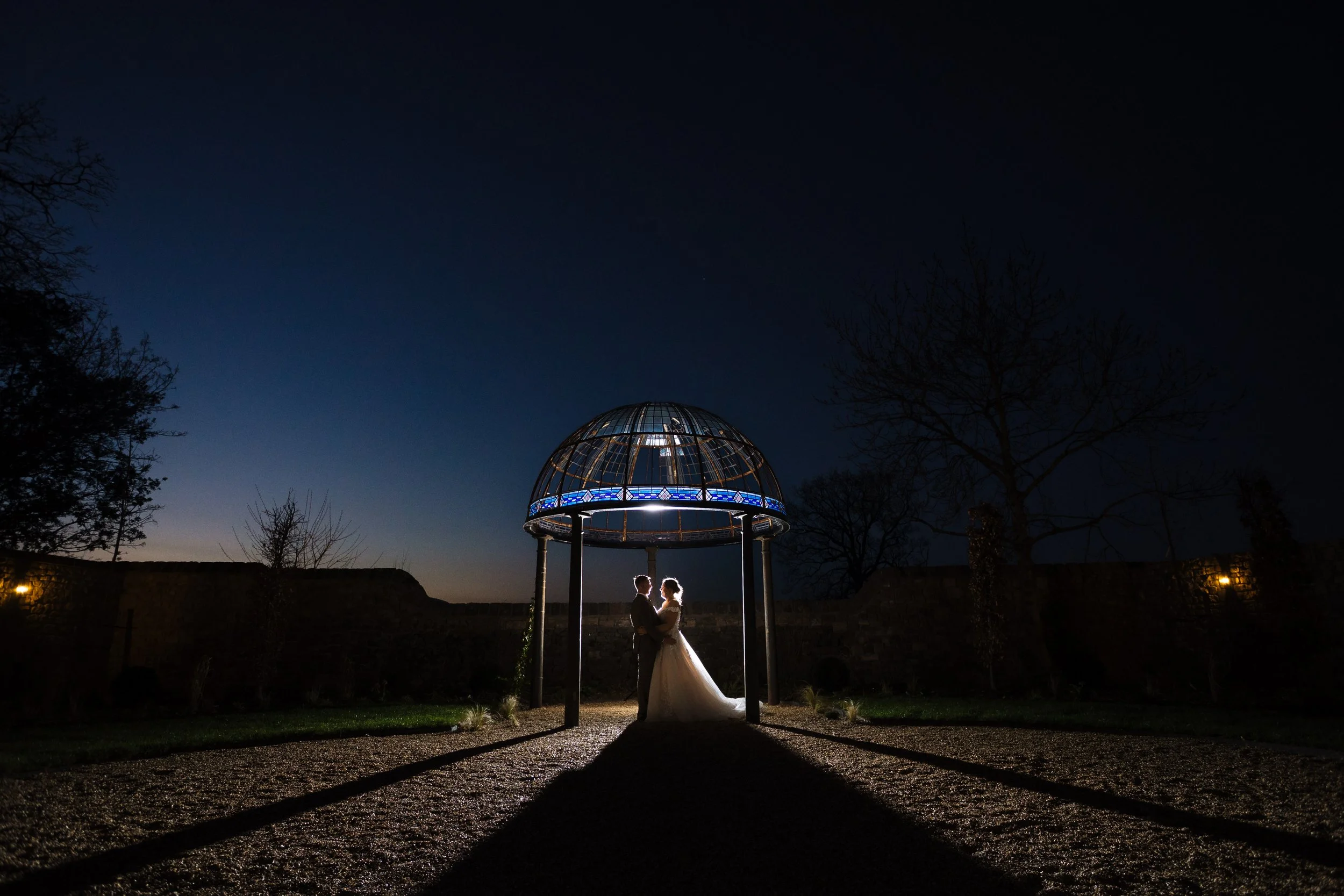 Couple in wedding attire standing under a metal gazebo at night, backlit to create silhouette, with a clear dark sky and sparse trees in the background.