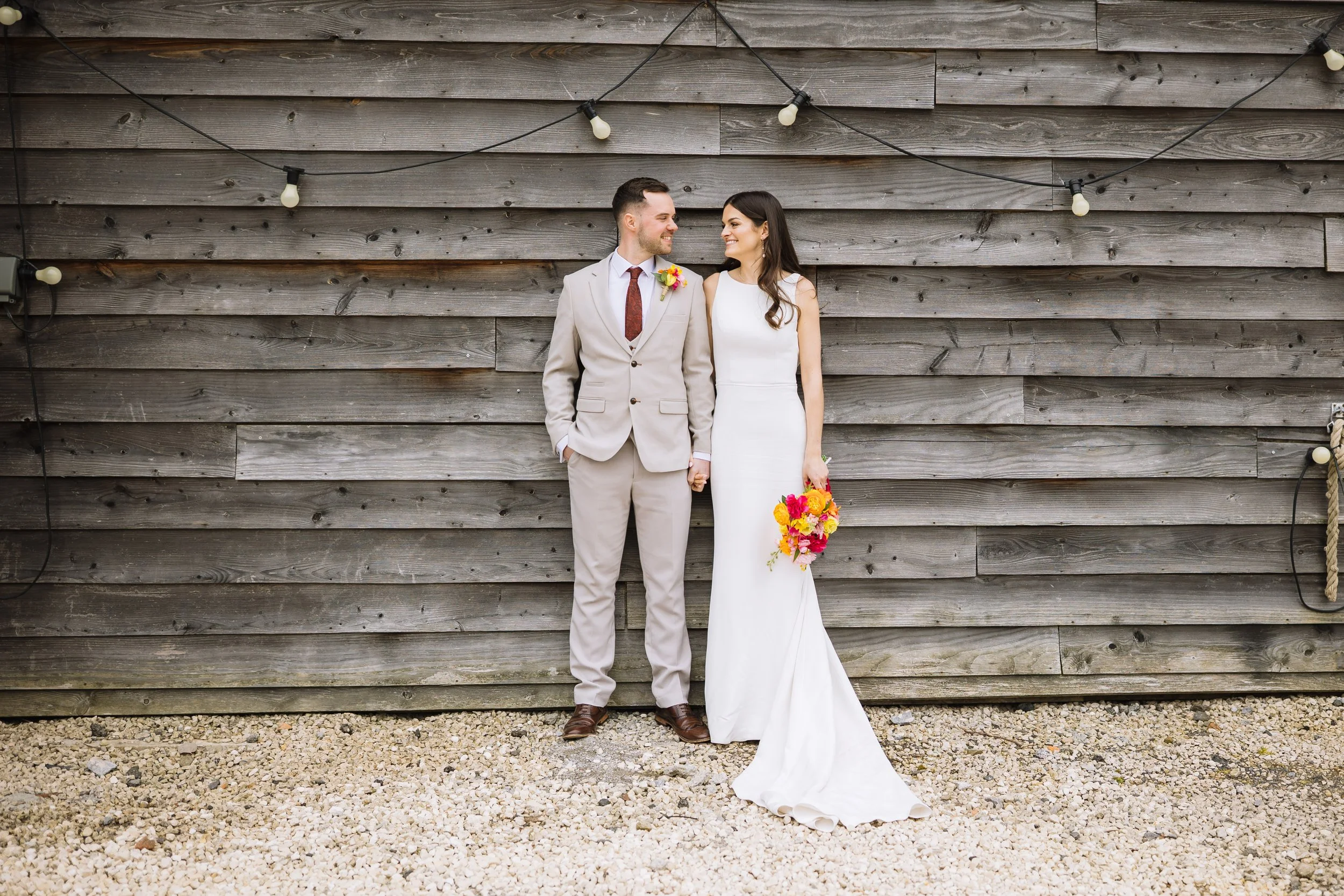 A couple in wedding attire standing against a rustic wooden wall decorated with string lights, holding hands and looking at each other. The bride is holding a colorful bouquet of flowers.