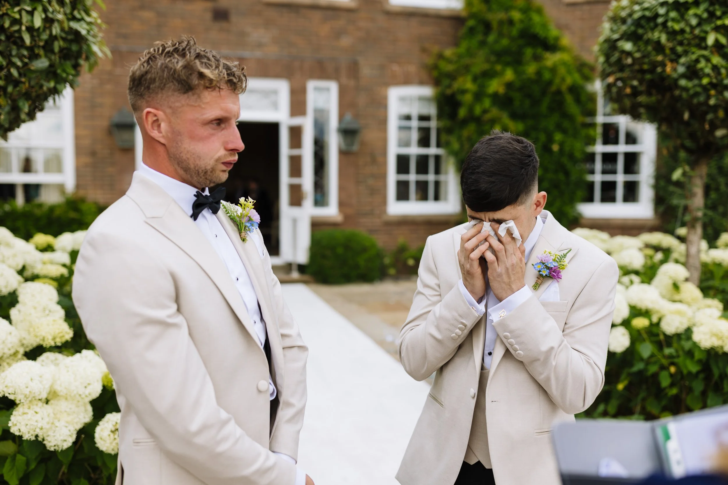 Two men in beige suits with boutonnieres on their lapels, standing outdoors near white flowers and green trees. One man is wiping tears from his eyes with tissues, while the other looks on somberly.