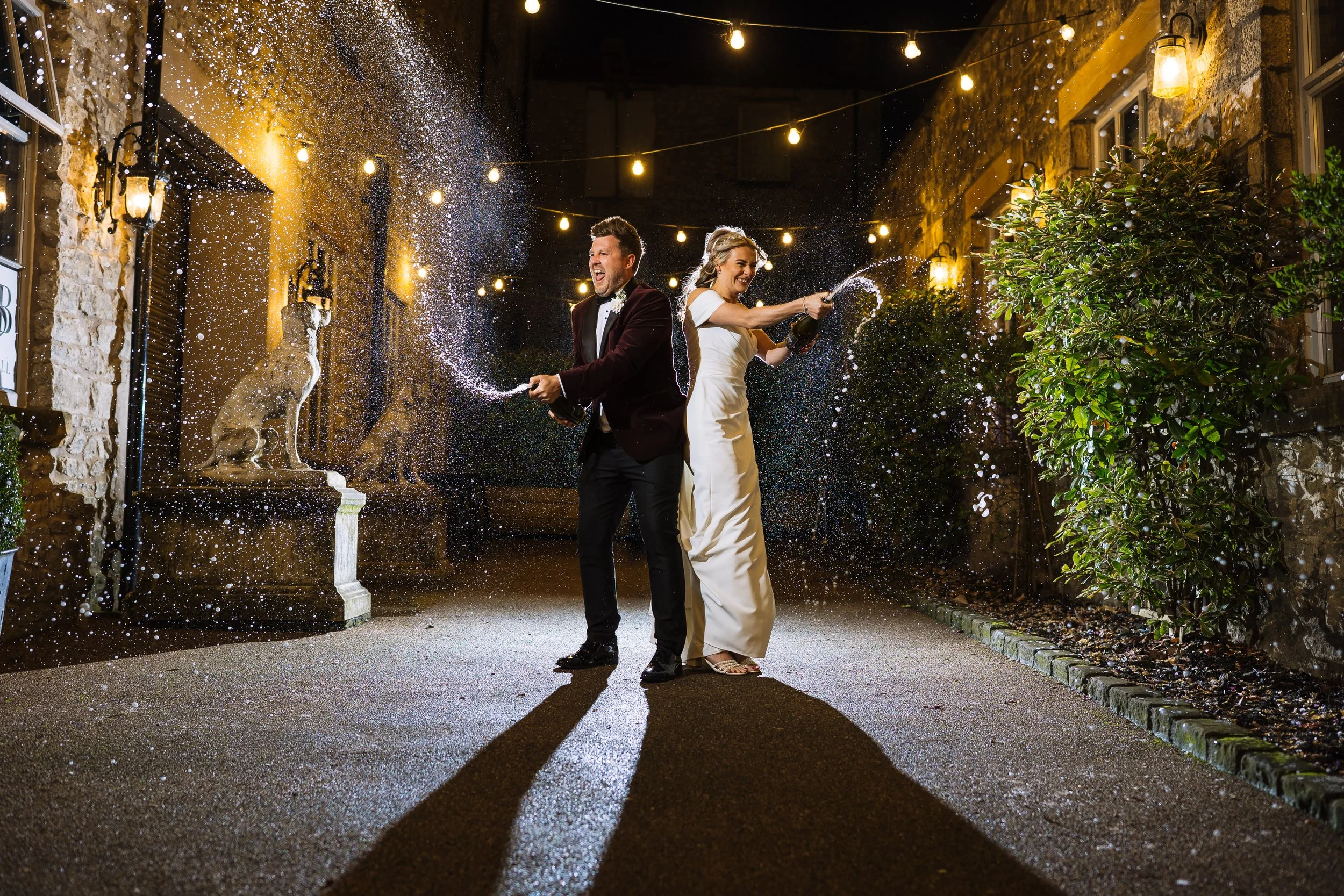 A newlywed couple celebrating at night with champagne, spraying it into the air. The scene is decorated with string lights and flanked by stone walls, with dogs in the background.