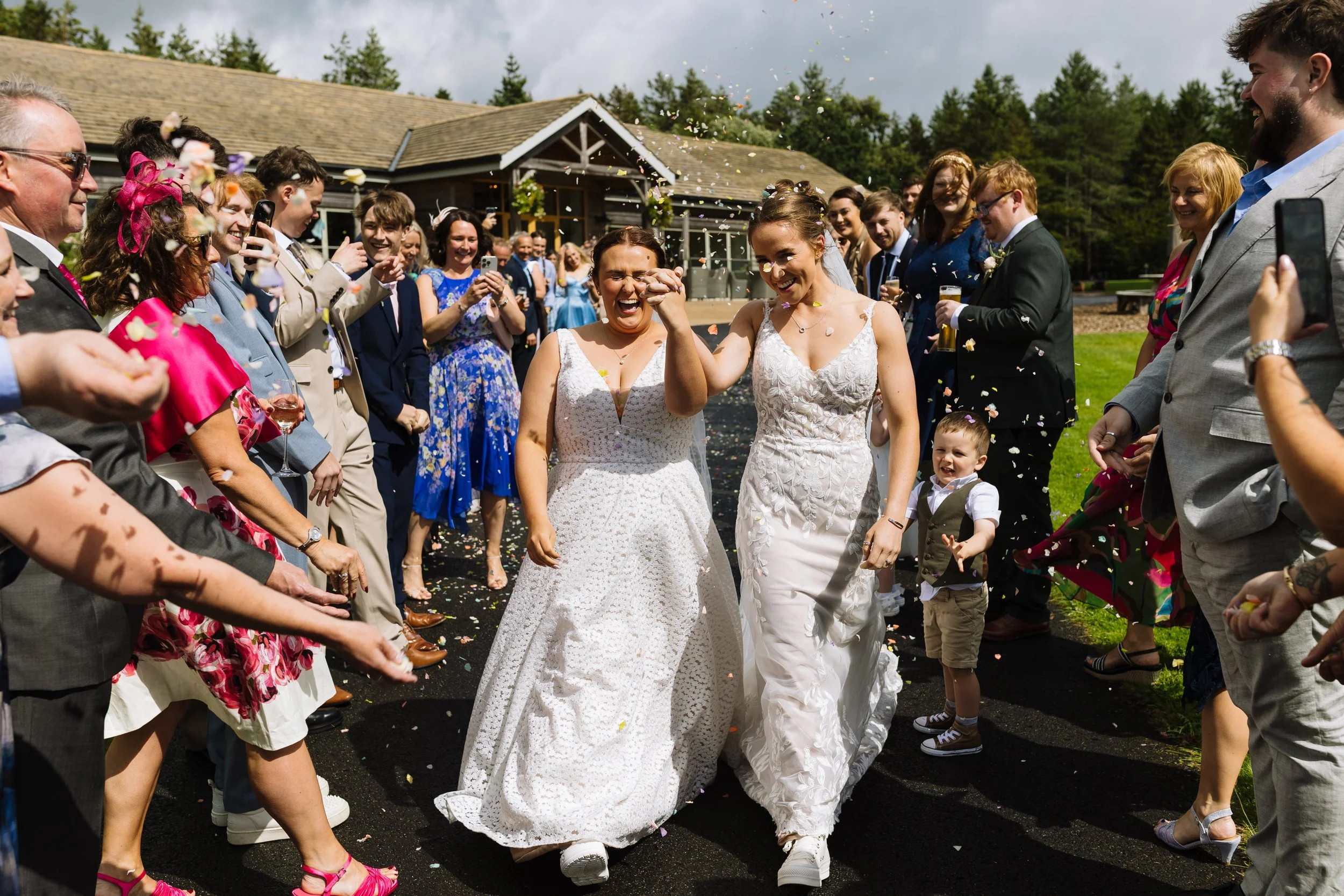 Two women, one with a short haircut and white dress, the other with long hair and a white wedding dress, smiling and holding hands as they walk through a crowd of people celebrating outside on a sunny day. Guests are cheering and taking pictures.
