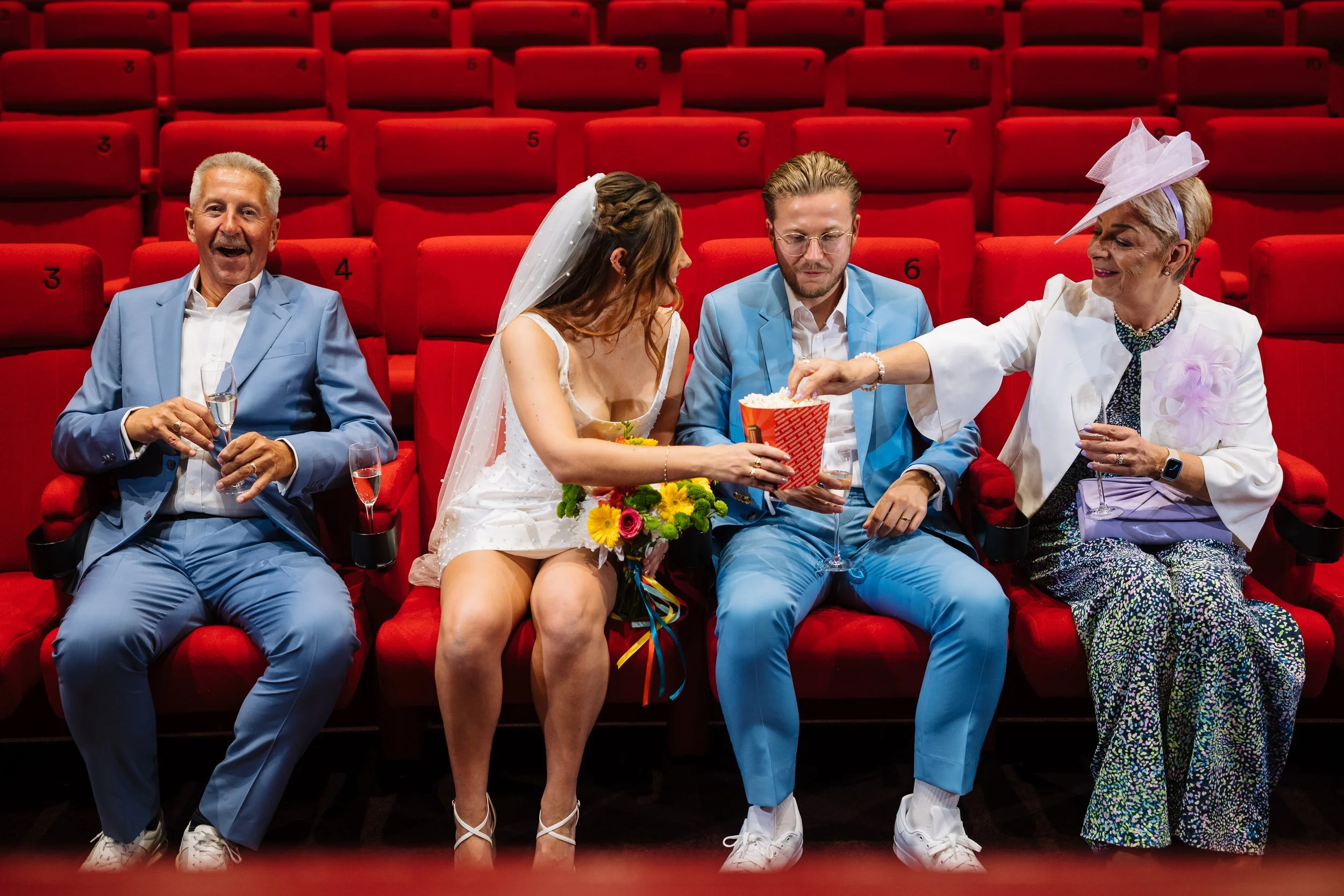 A group of five people sitting in red theater seats, dressed in formal and colorful attire, with two people giving popcorn to a bride and groom. The bride is in a white dress with a veil, and the groom is in a light blue suit. Two older women are on 