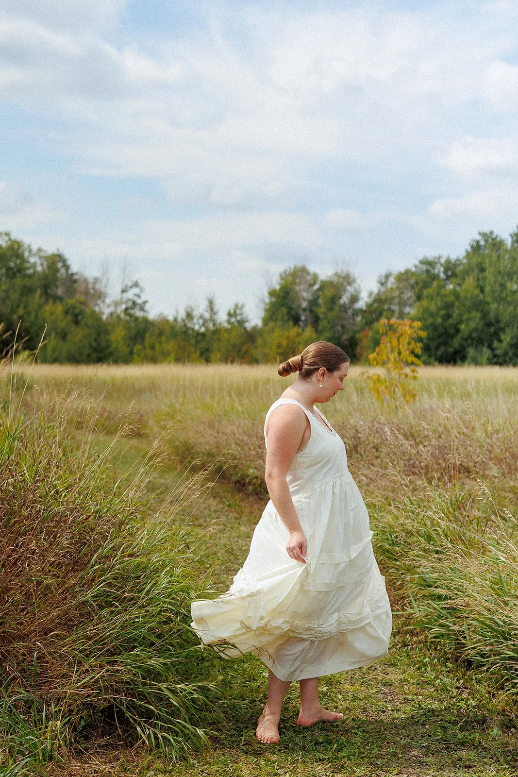 A pregnant woman in a white dress walking barefoot through a grassy field, looking down at her dress.