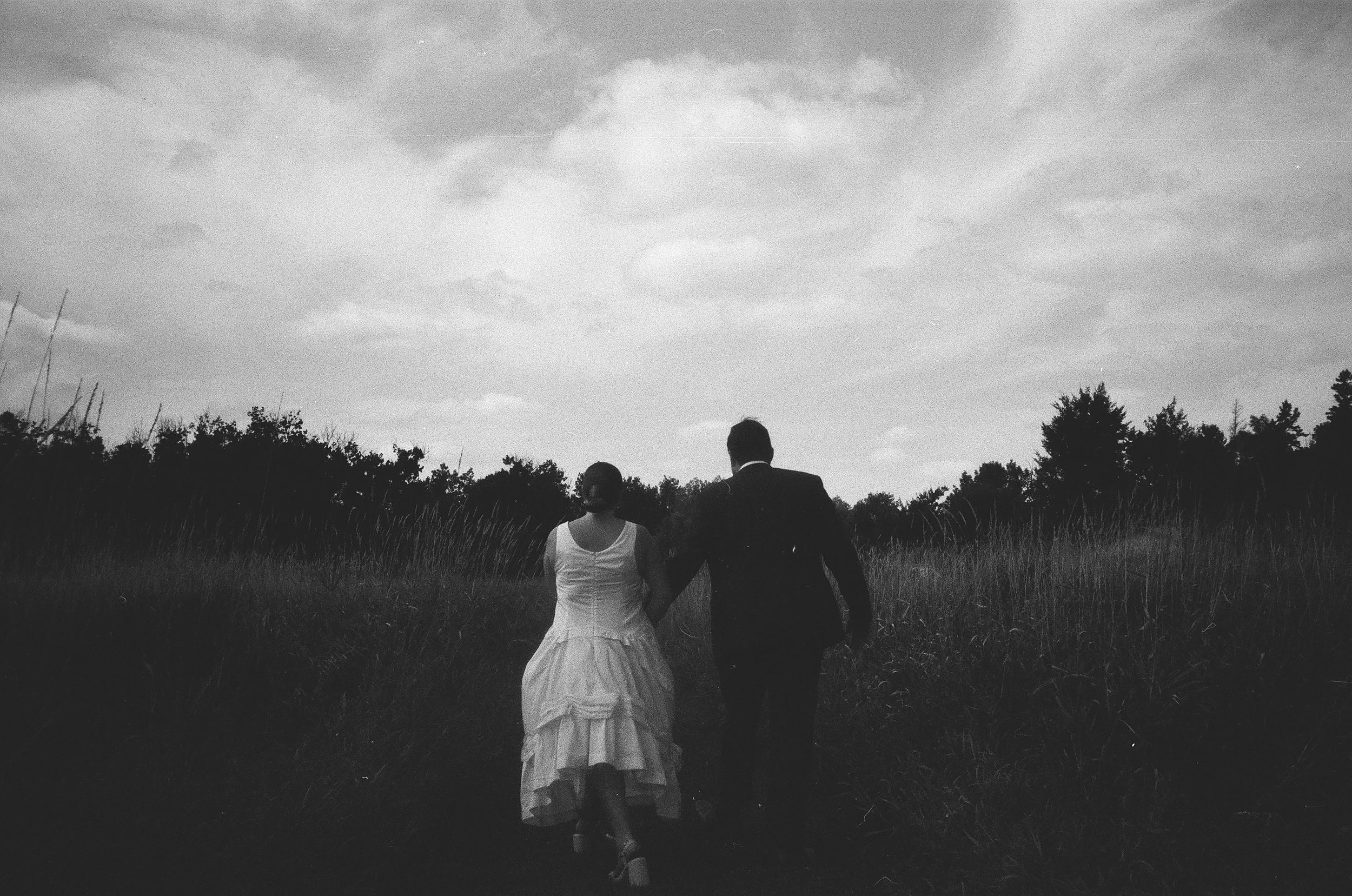 A black and white photo of a man and woman walking hand-in-hand through a field, with trees in the distance and a cloudy sky overhead.