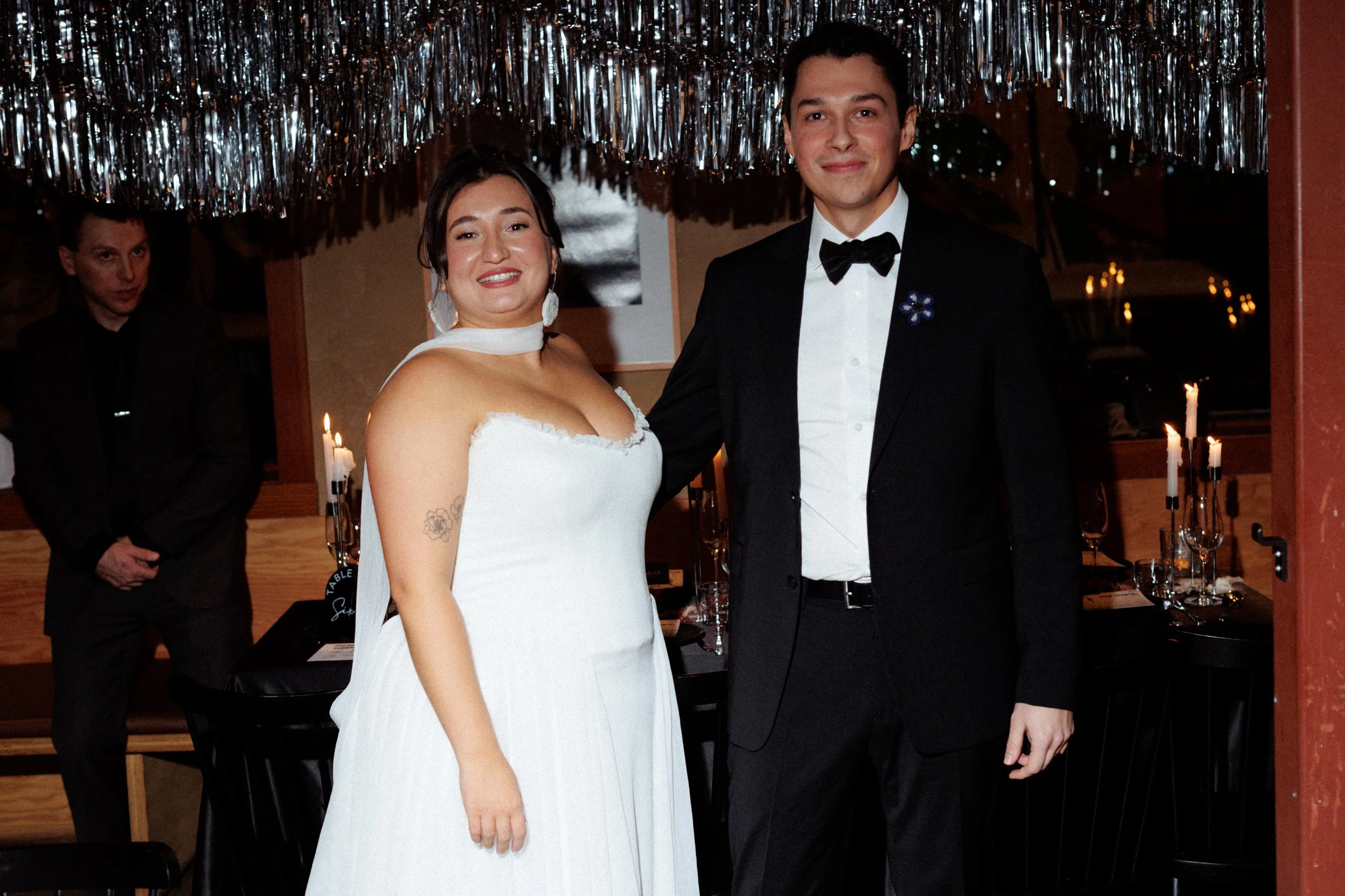 A bride and groom standing together at their wedding reception, smiling at the camera. The bride wears a white strapless wedding gown, while the groom wears a black tuxedo with a bow tie. They are indoors with decorative silver streamers hanging from