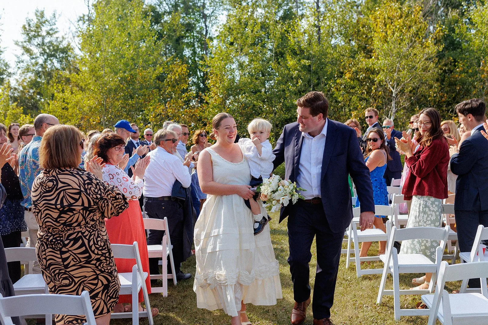 A newly married couple walking down the aisle outdoors, surrounded by guests clapping and celebrating, with trees and a bright sky in the background.