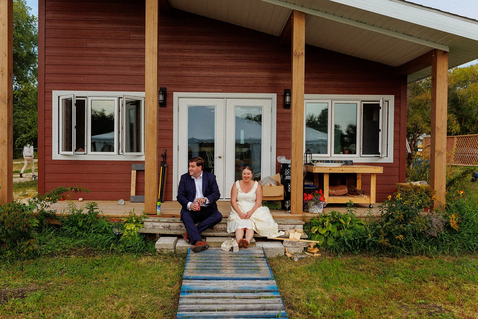 A man and woman sitting on the steps of a red house with a porch, amidst a garden with green plants and flowers. The man is wearing a dark suit and holding a drink, while the woman is in a white dress smiling. There are construction materials and a w