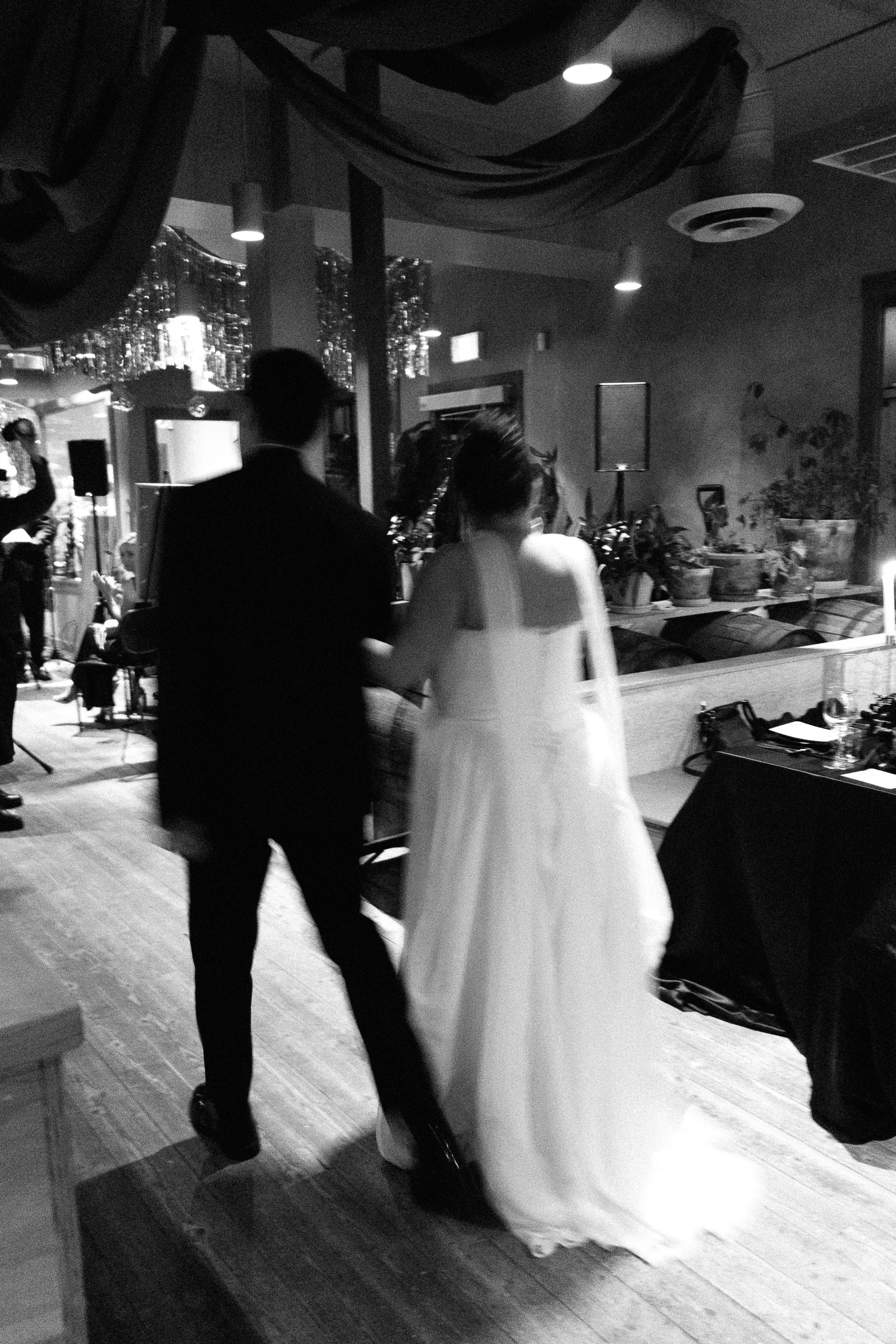 A black and white photo of a bride and groom walking together in a dimly-lit reception space, with a band playing in the background and plants and decorations on the side.