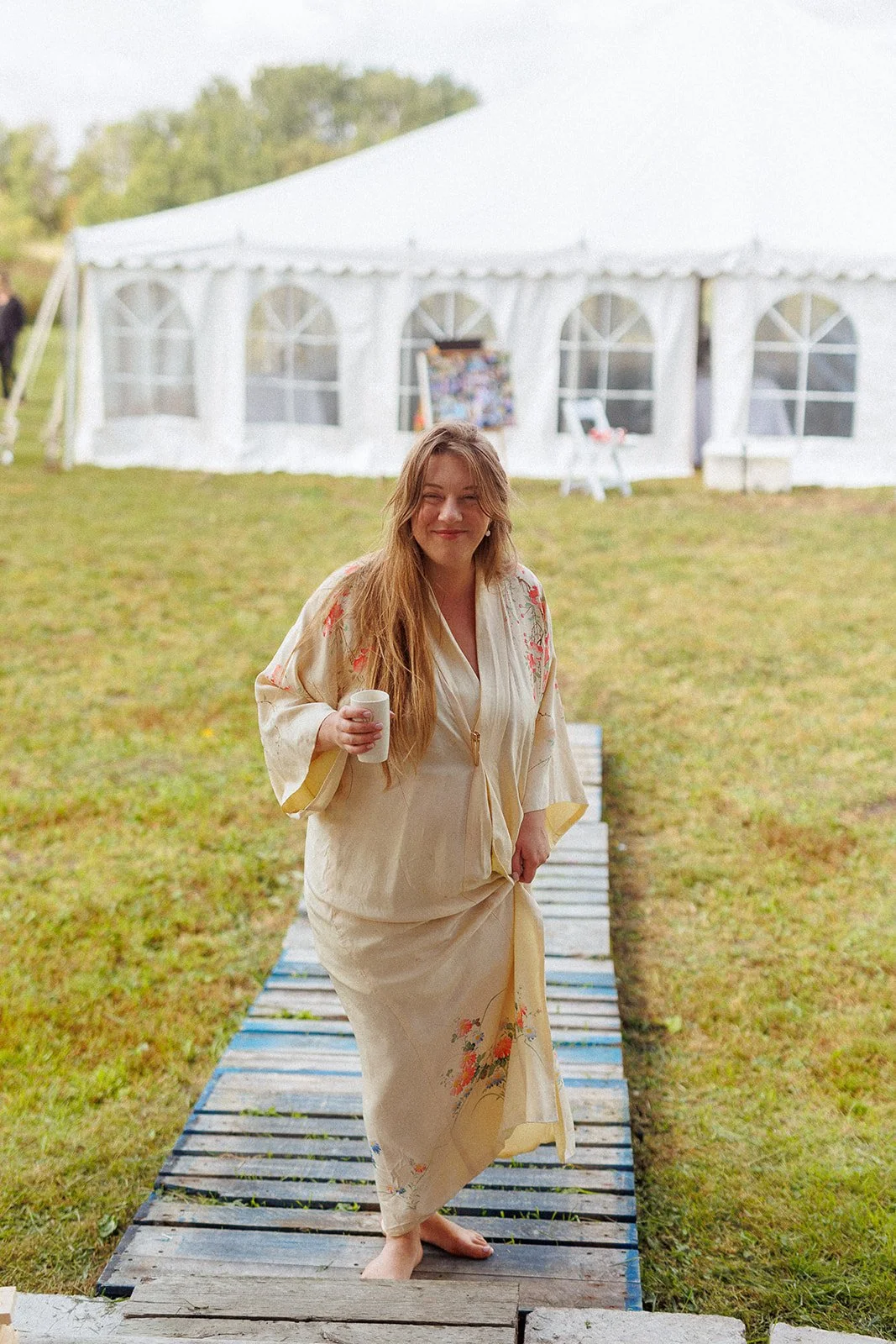 Woman in a cream-colored floral kimono holding a white mug walking on a wooden path outdoors in front of a white event tent.