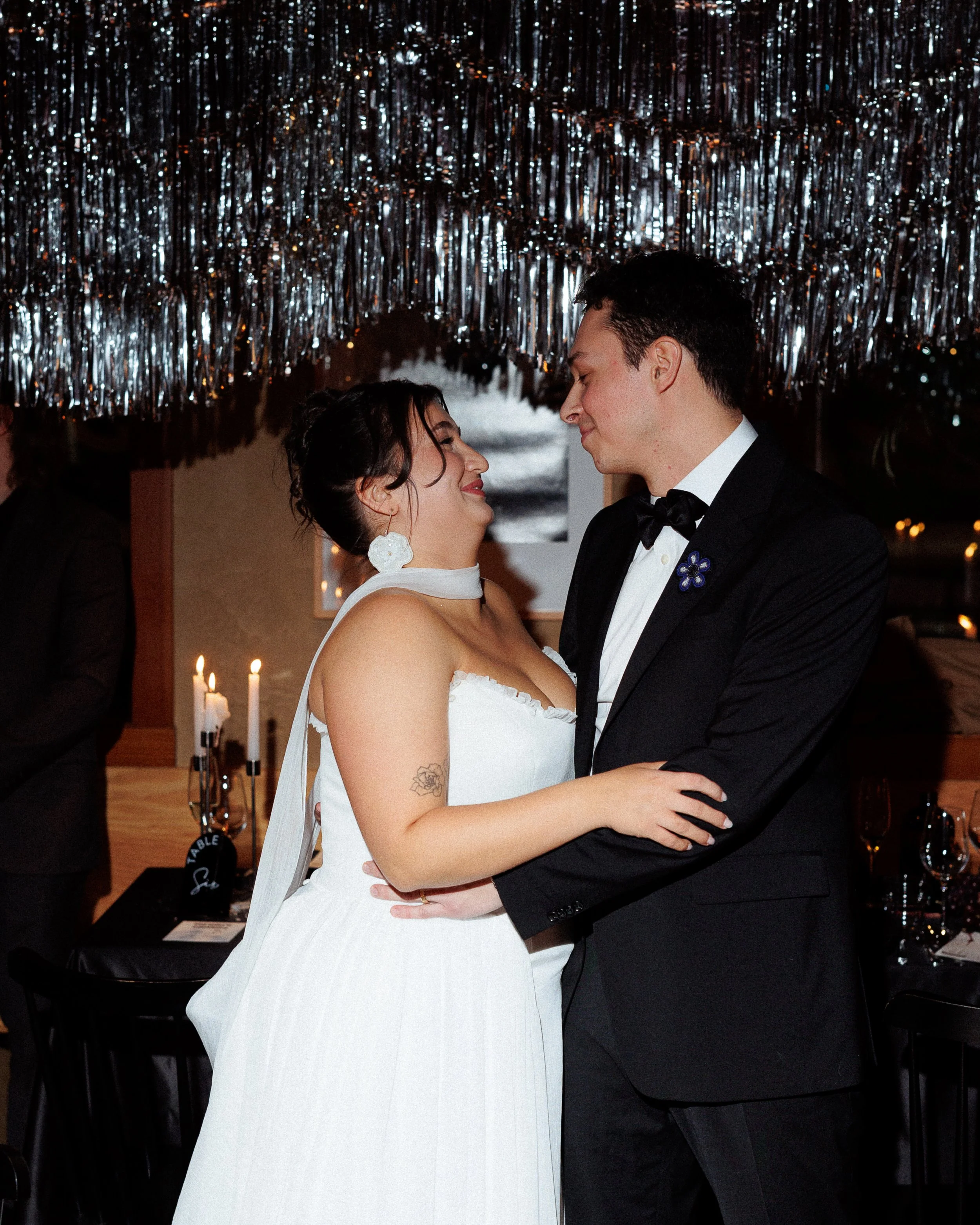 A couple dressed in formal attire, the woman in a white dress and the man in a black tuxedo, sharing a dance at a wedding reception with a shiny silver decoration overhead.