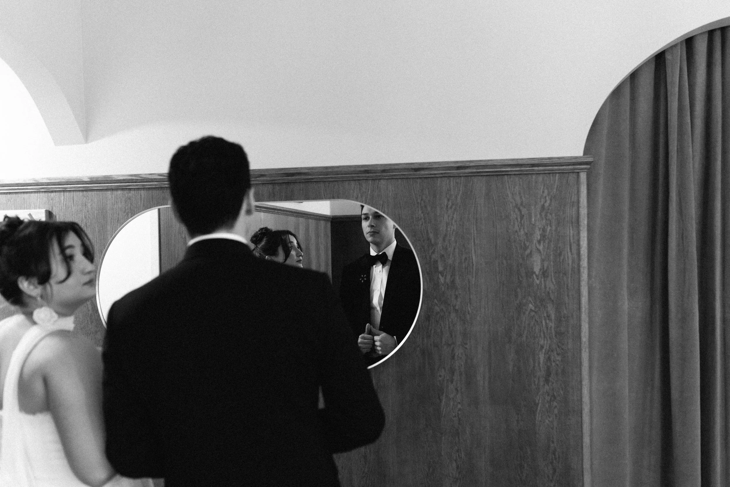 A black and white photo of a man in a tuxedo looking into a mirror, with two women nearby, in a room with wooden wall paneling and a curtain.