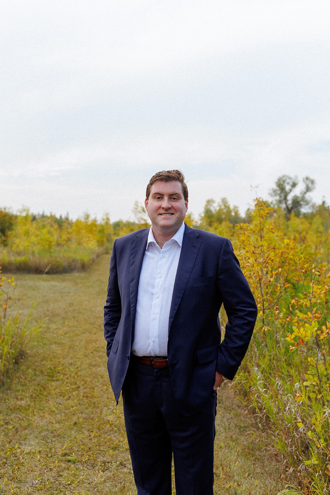 A man in a navy blue suit and white shirt standing outdoors on a grassy path, with trees and bushes showing autumn foliage in the background.