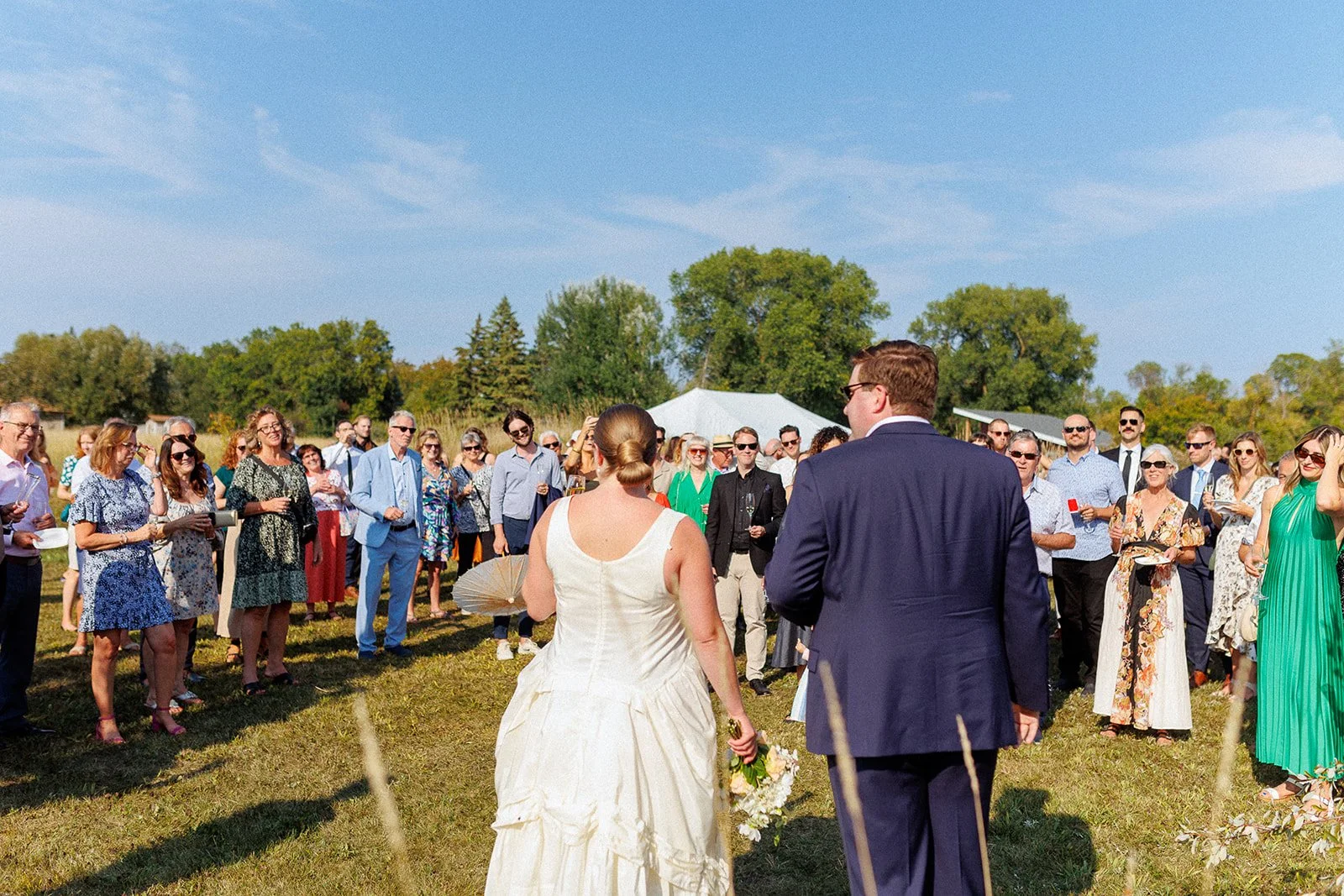 A wedding ceremony outdoors on a sunny day with a bride and groom standing in front of a group of guests, green trees in the background, and a clear blue sky.