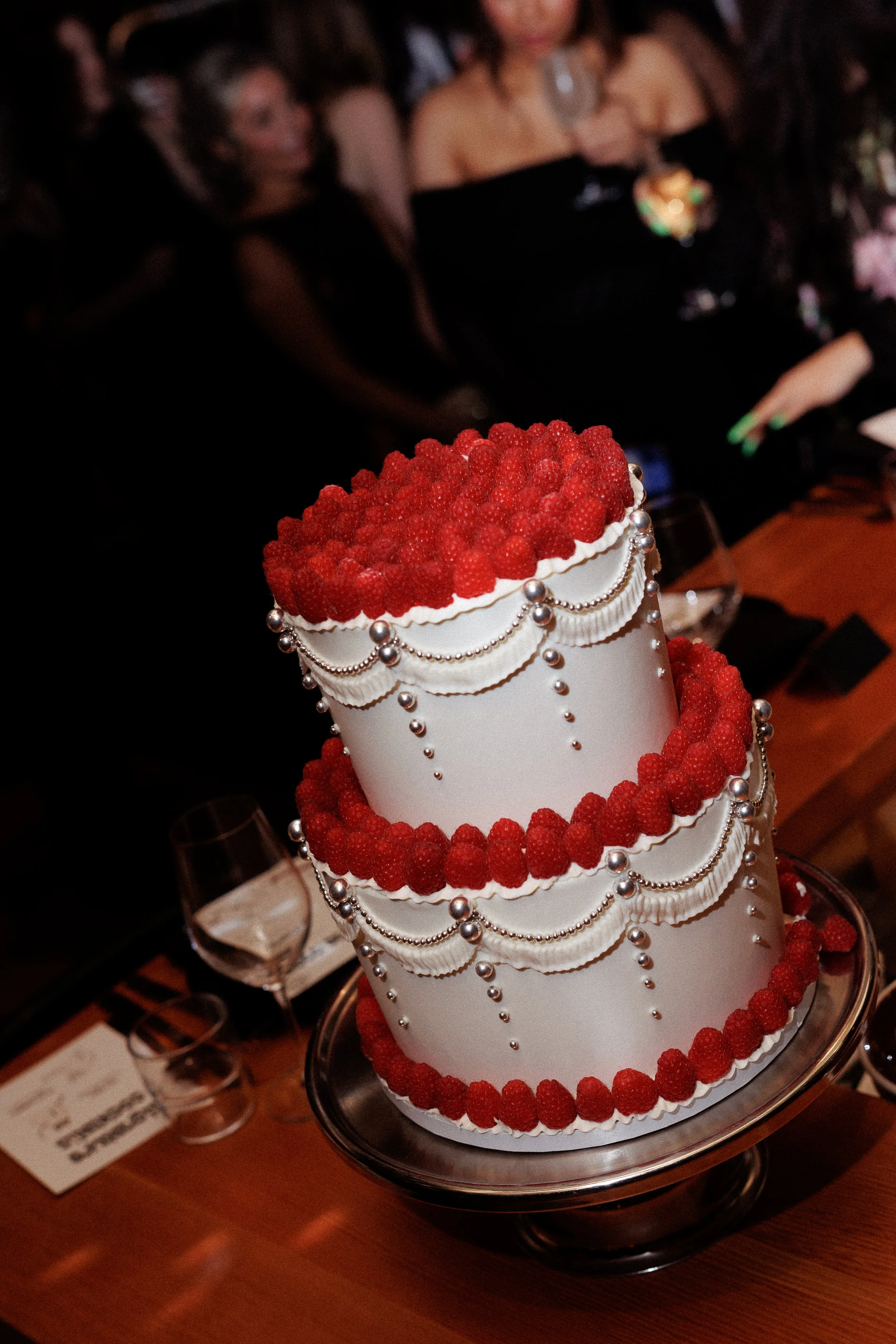 A three-tiered white cake decorated with raspberries and silver beads, displayed on a cake stand at a celebration event.