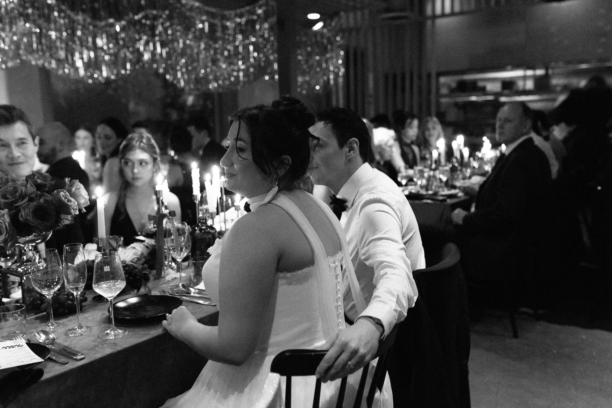 Black and white photo of a dinner party with multiple guests seated at a long table, decorated with candles and flowers, in a dimly lit room.