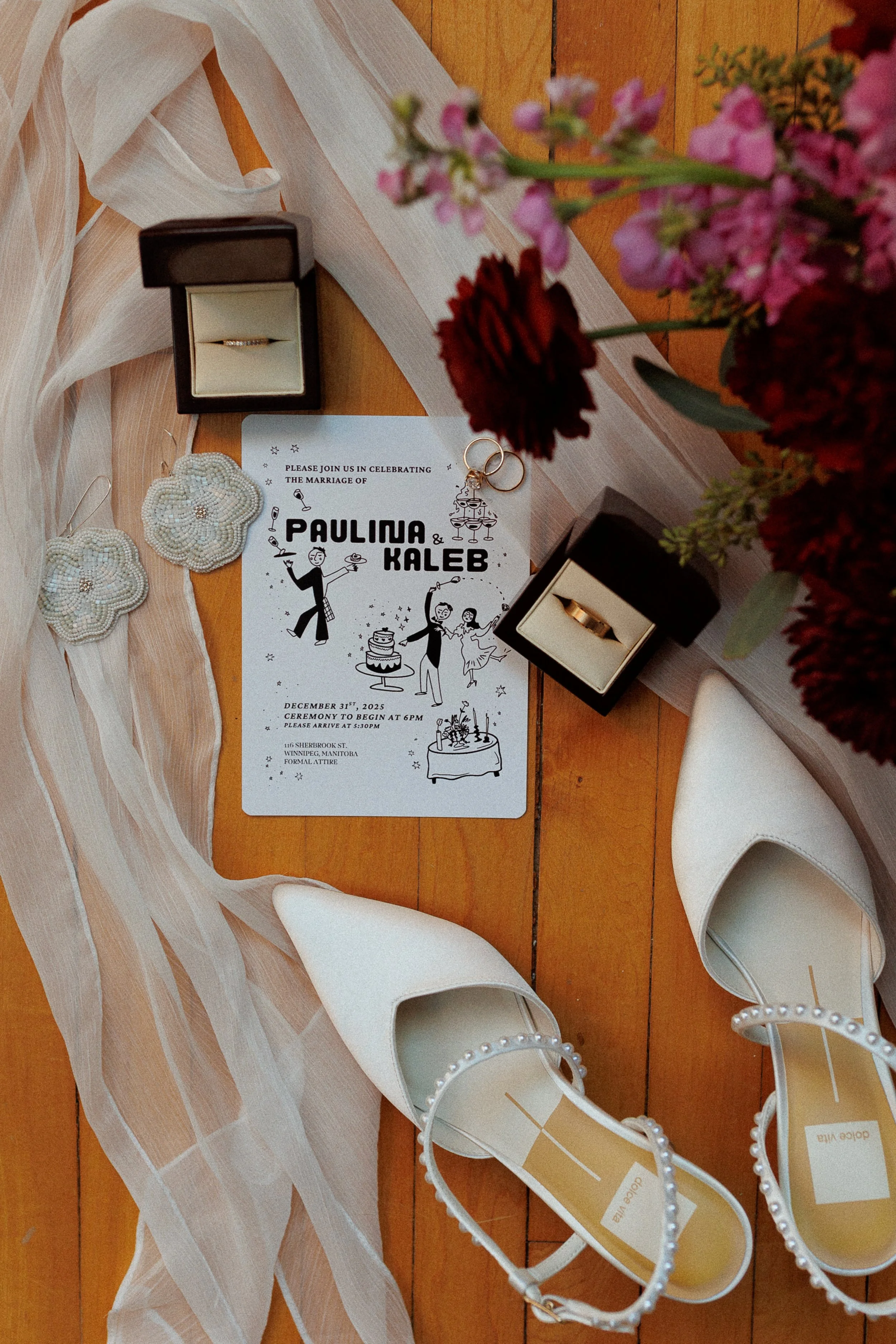 Wedding invitation, white shoes with pearl ankle straps, a black ring box with a gold wedding band, a bouquet of red and pink flowers, and beaded flower-shaped earrings laid on a wooden floor with a sheer white fabric.