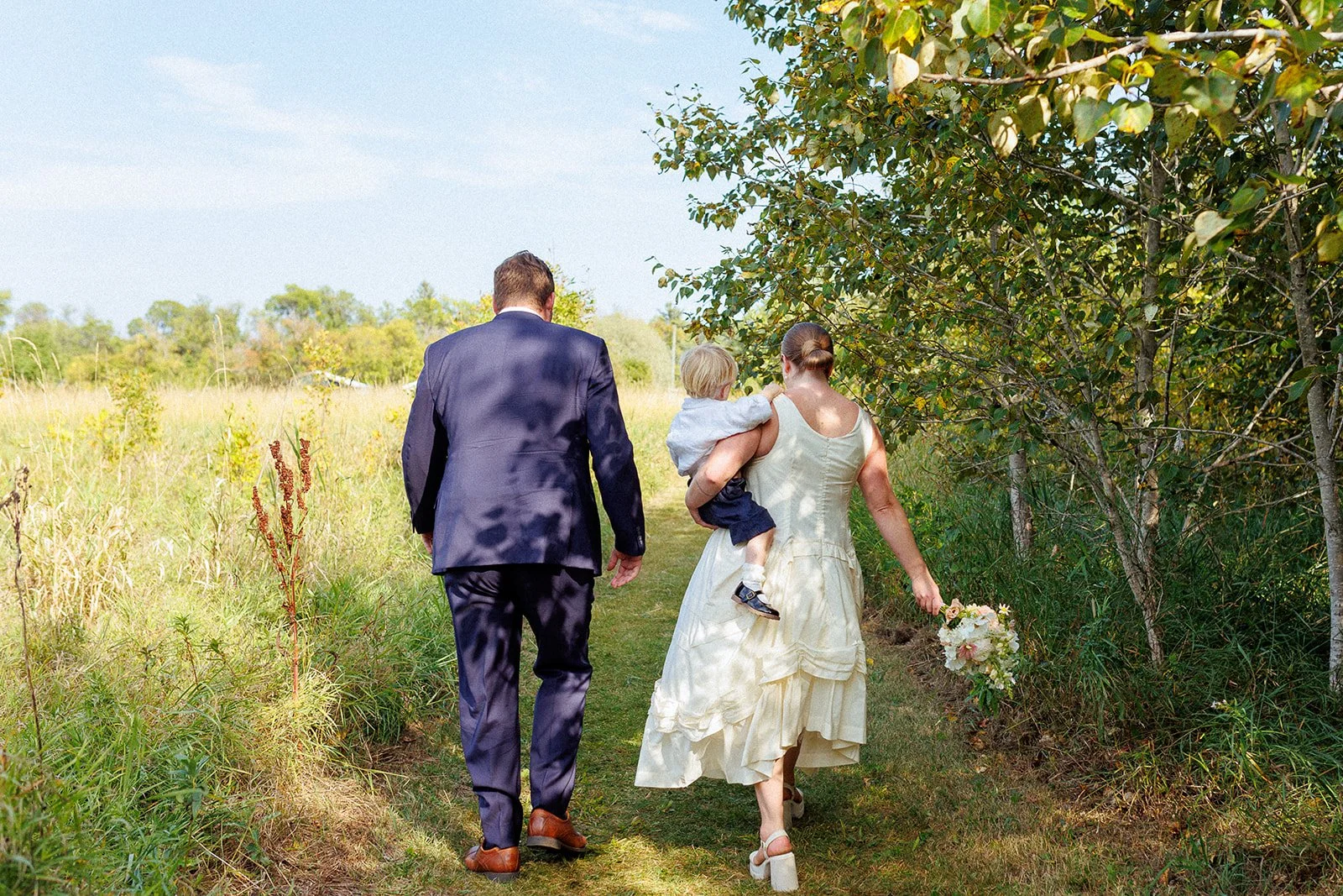 A couple with a small child walking on a grassy trail in a natural outdoor setting. The woman is holding a bouquet, and the child is resting an arm on her shoulder. The man is dressed in a dark suit, and the woman in a white dress. They are walking a