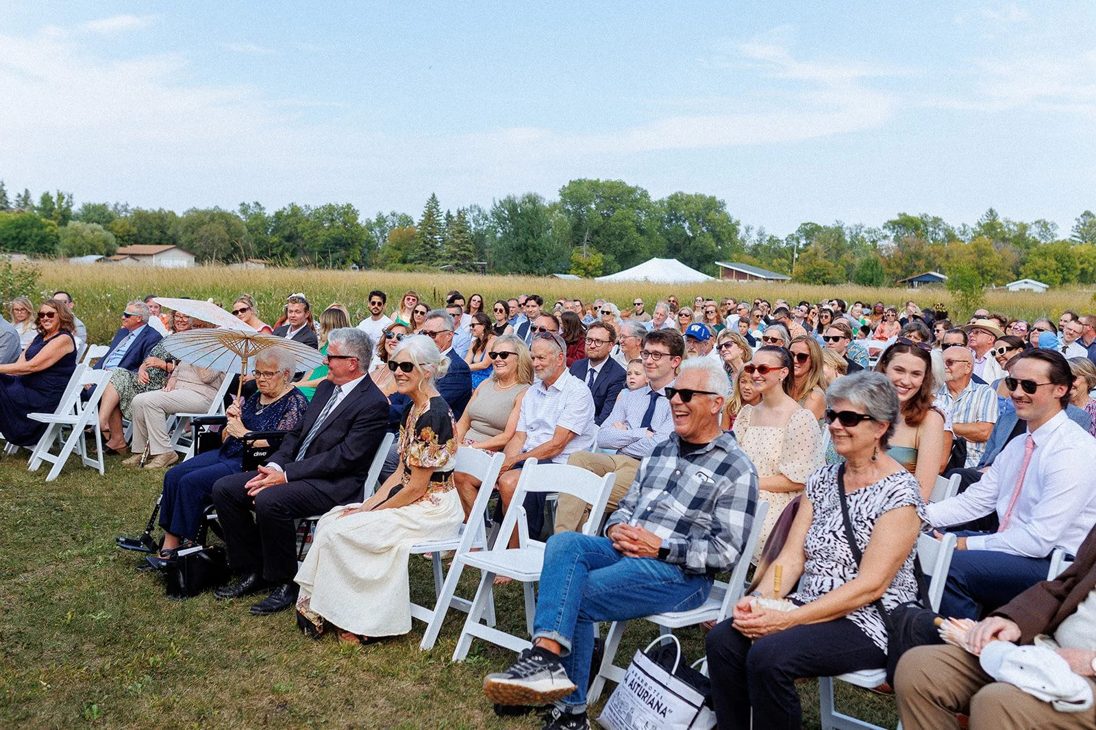 Crowd of people attending an outdoor event on a grassy field, seated on white chairs, with many smiling and enjoying the event on a sunny day.