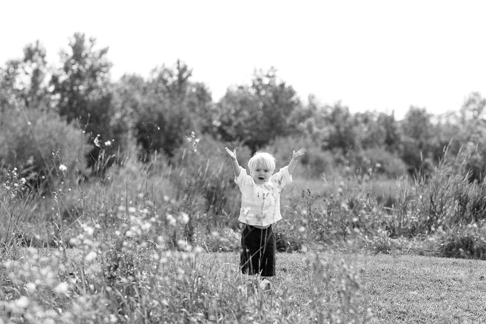 Black and white photo of a young child with arms raised, standing in a grassy field with trees in the background.