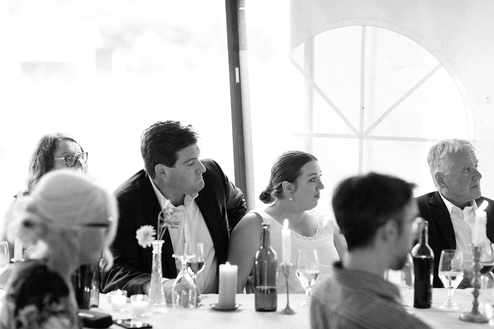 Group of people sitting at a table during a formal event, with candles and wine glasses, in front of a large window with a decorative arch.