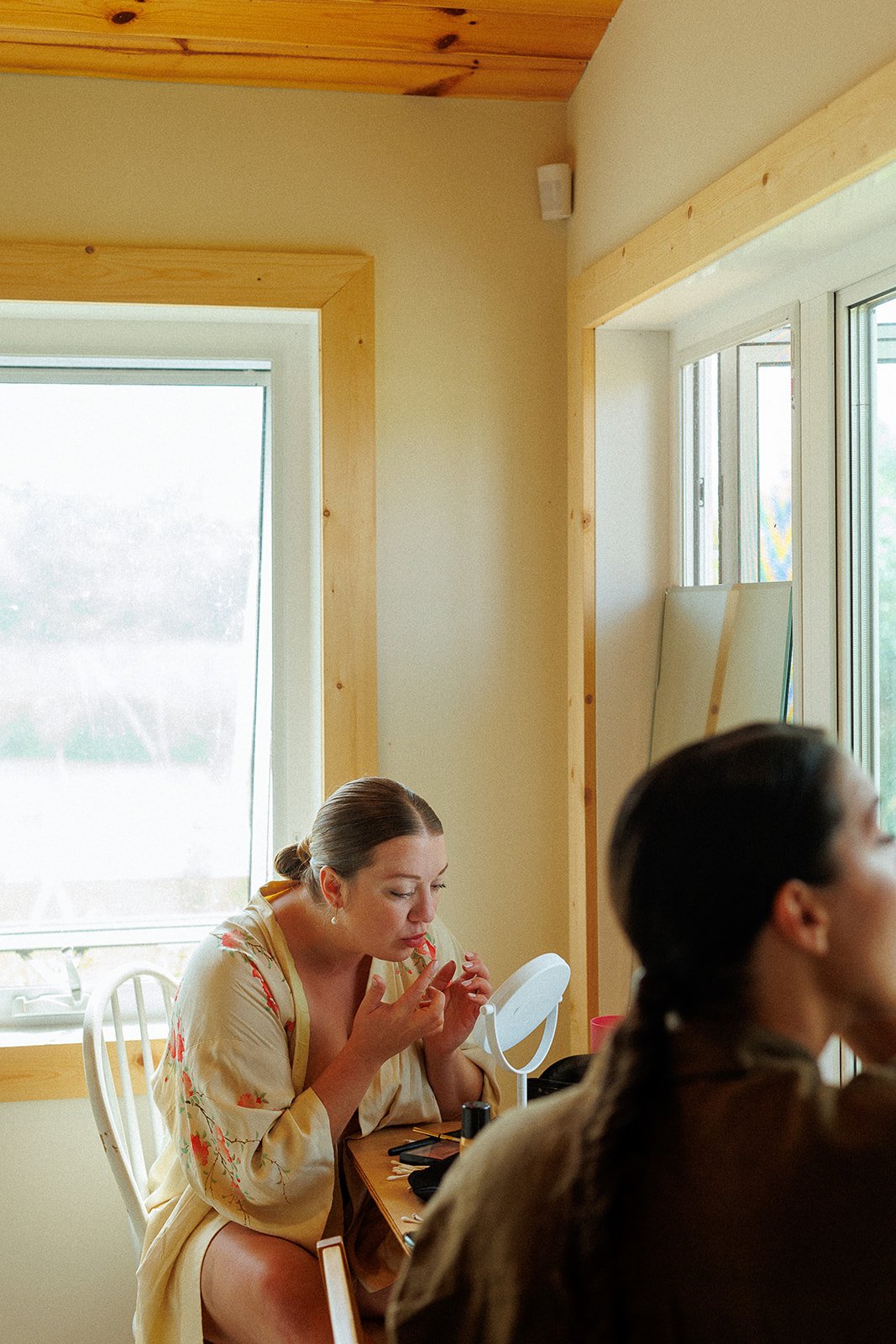 A woman in a floral robe applies makeup in front of a small mirror while sitting at a wooden desk near a window.