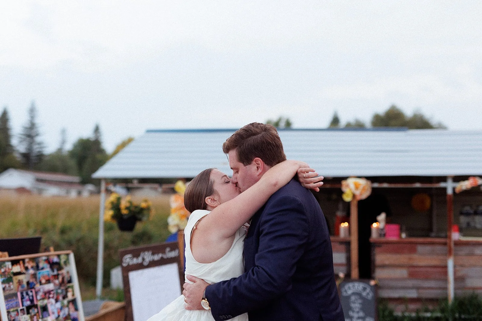 A couple kissing outdoors at a wedding reception, with a decorated wooden stand and photo display in the background.