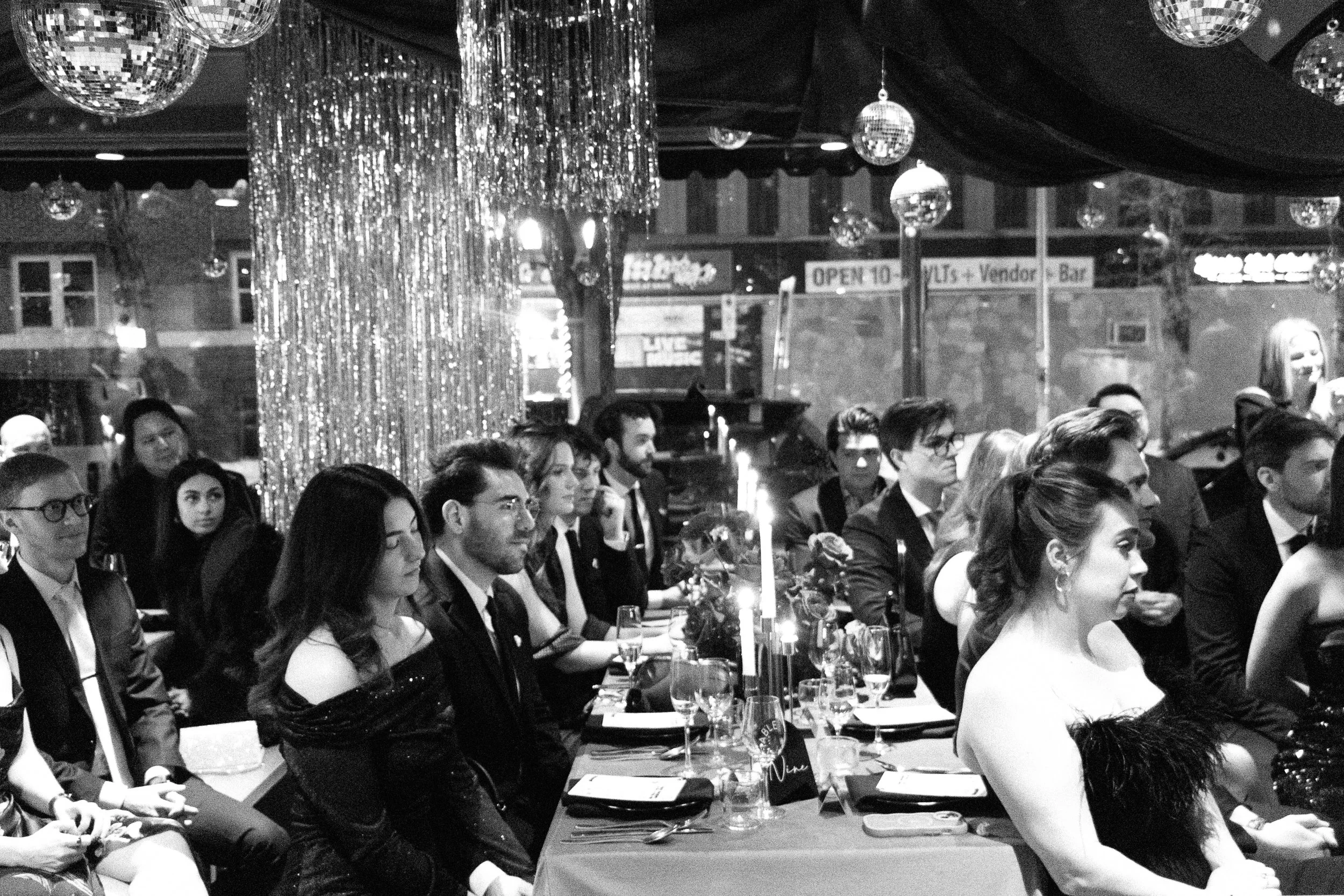 A black and white photograph captures a formal dinner event in a decorated venue. Attendees, dressed in elegant attire including suits and evening dresses, are seated at tables adorned with candles, glasses, and menus. The background features shiny h
