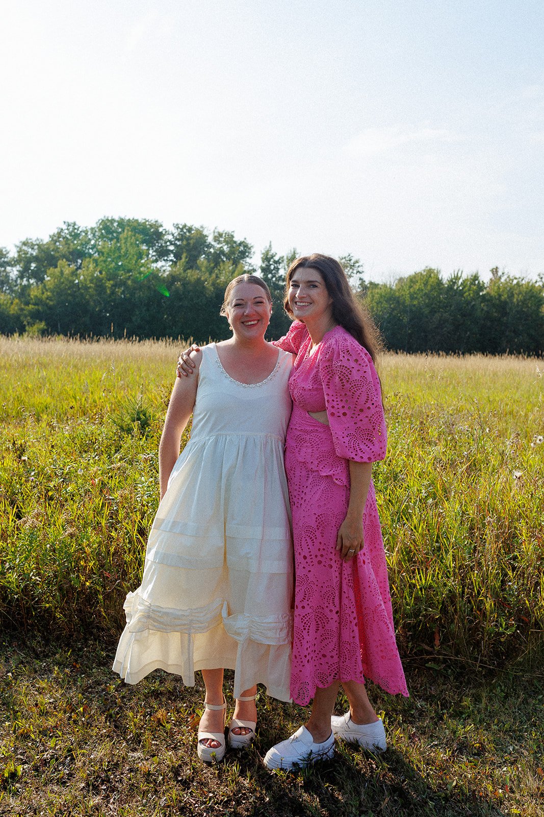 Two women standing together in a field, smiling, one in a white dress and the other in a pink dress.