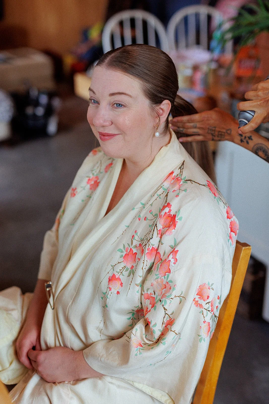 Woman with dark brown hair in a low ponytail, wearing a cream silk robe with pink and orange floral patterns, getting her hair styled with a curling iron by a stylist with tattoos.