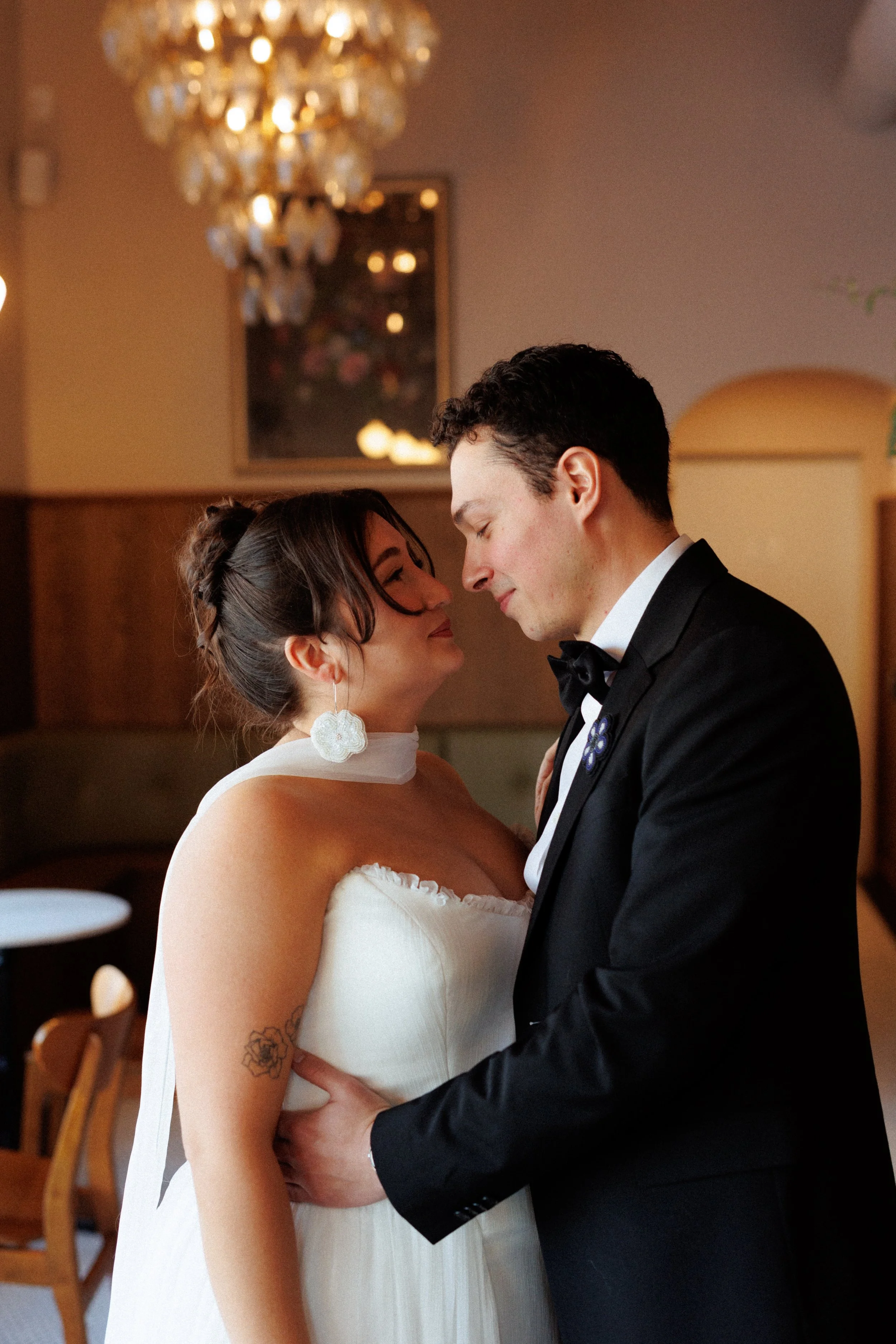 A bride and groom sharing a tender moment indoors, with the groom in a black tuxedo and the bride in a white strapless wedding dress, as they lean their foreheads together.