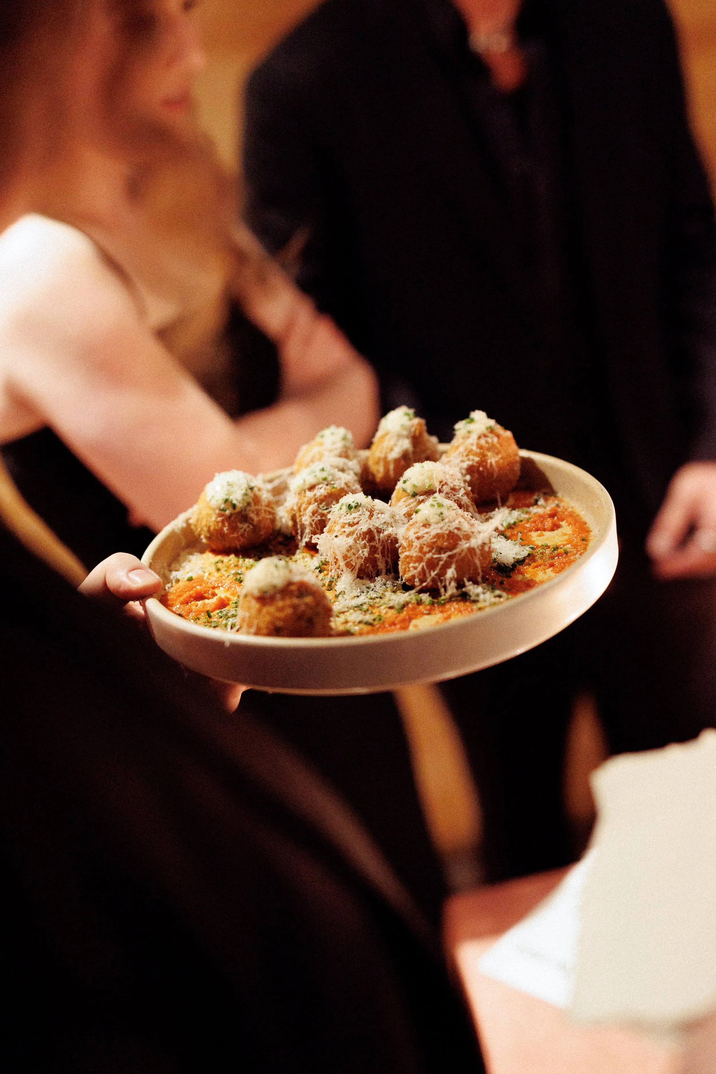 Person holding a plate of breaded appetizers on a bed of vegetables, with indistinct people in the background, at a social gathering or dinner event.