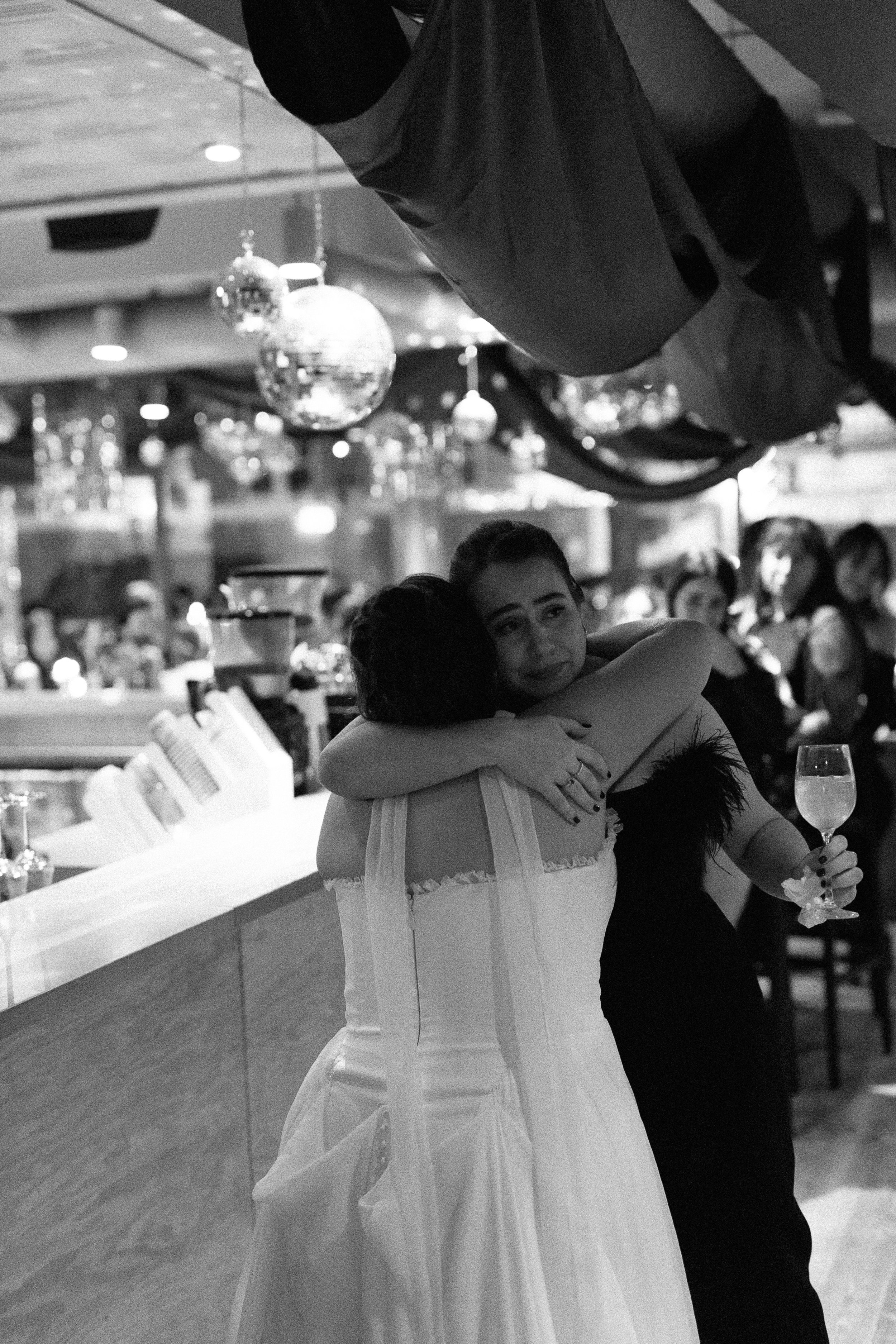 A woman in a white dress hugging another woman in a black dress at a celebration, with other guests and disco balls in the background.