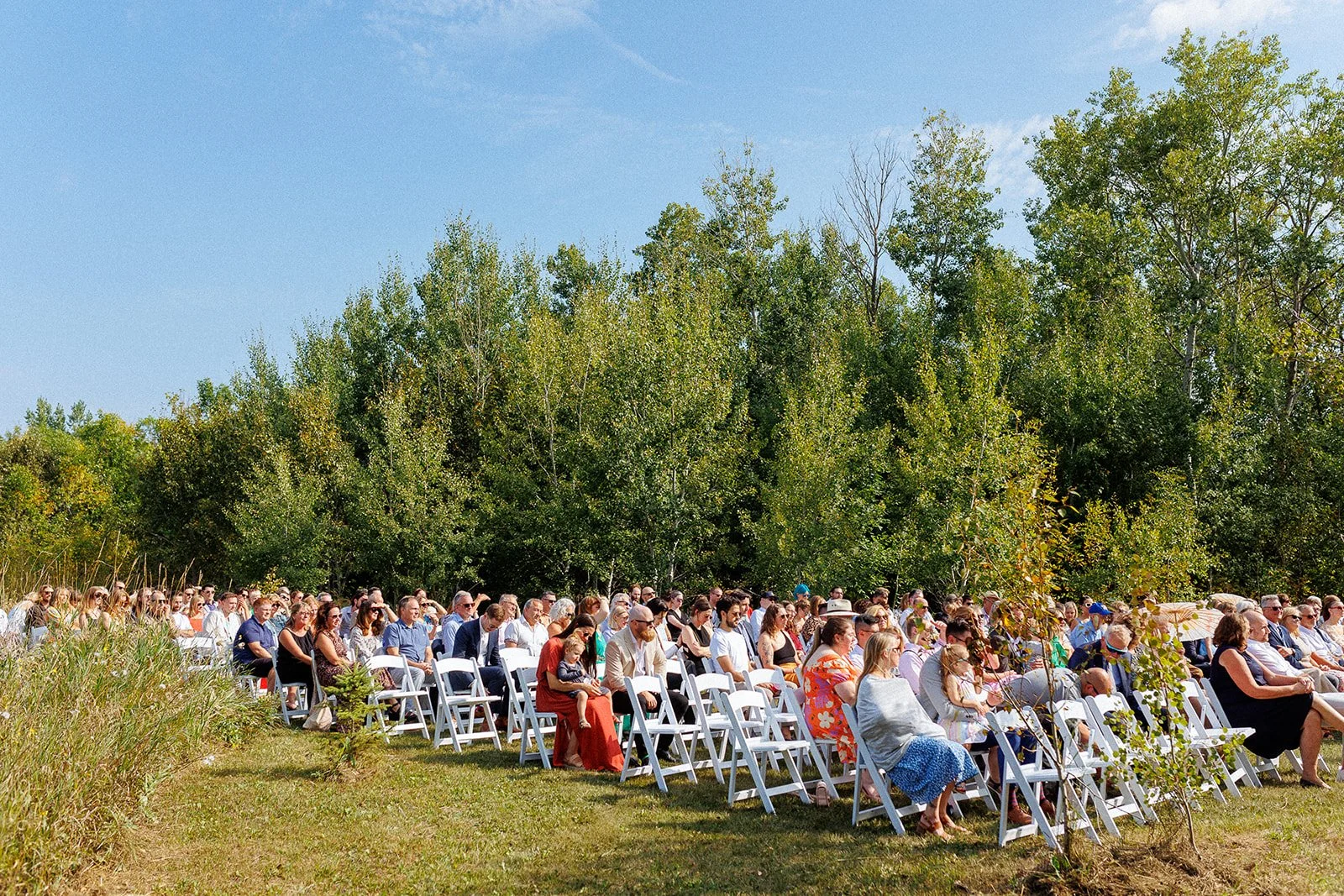 Group of people sitting on white chairs outdoors, attending an event on a sunny day with green trees and blue sky in the background.