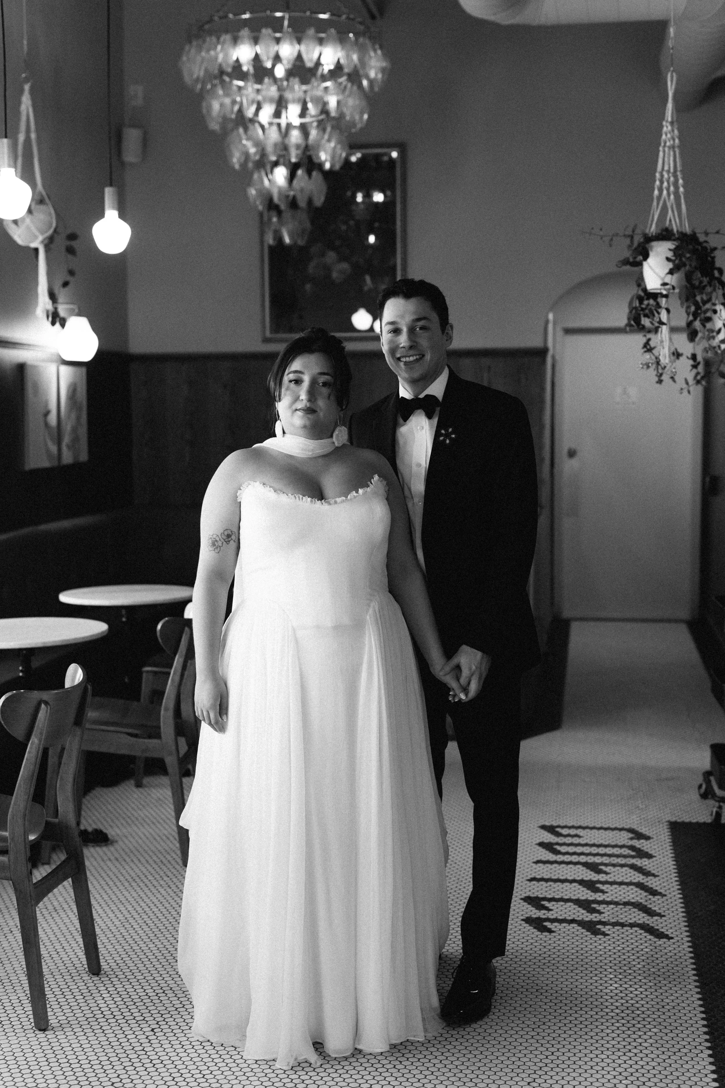 A bride and groom in wedding attire holding hands, standing inside a decorated venue with hanging lights and a chandelier.