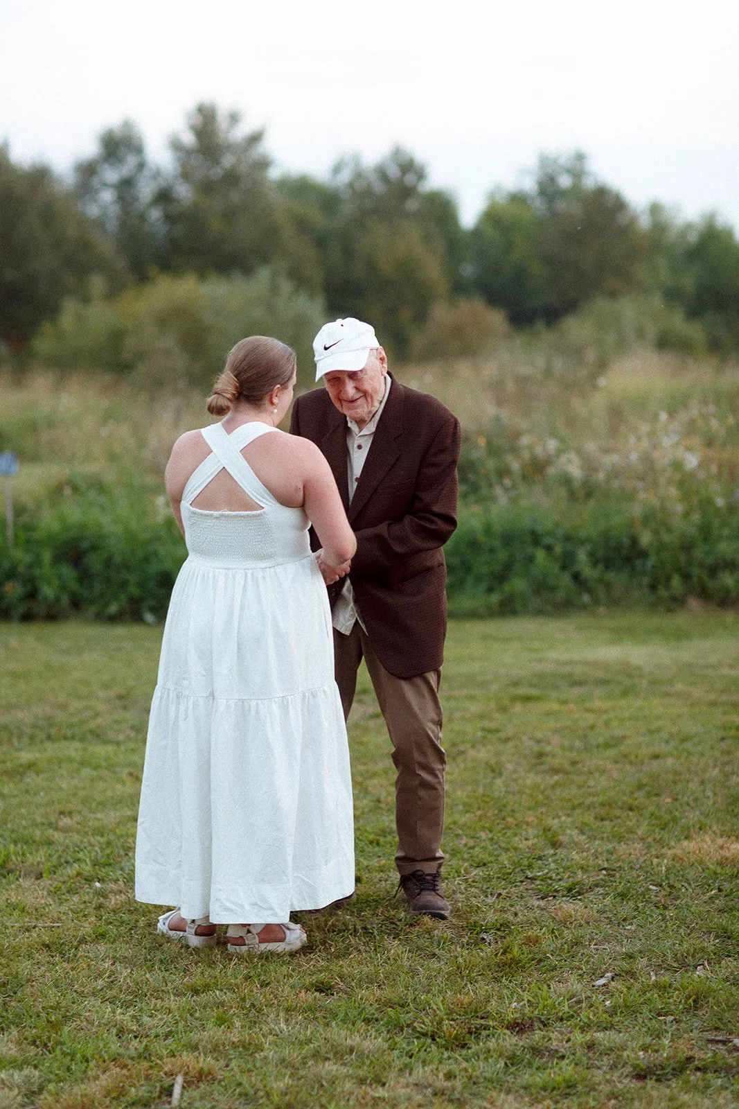 An elderly man in a brown blazer, khaki pants, and a white Nike cap holding the hands of a young woman in a white dress outdoors on a grassy field during daytime.