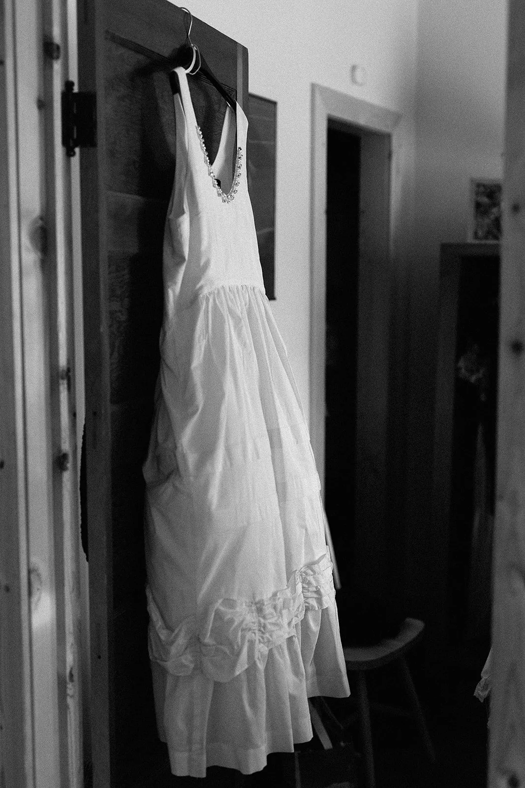 A white wedding dress hanging on a door with a beaded necklace draped over it, in a black-and-white photo.
