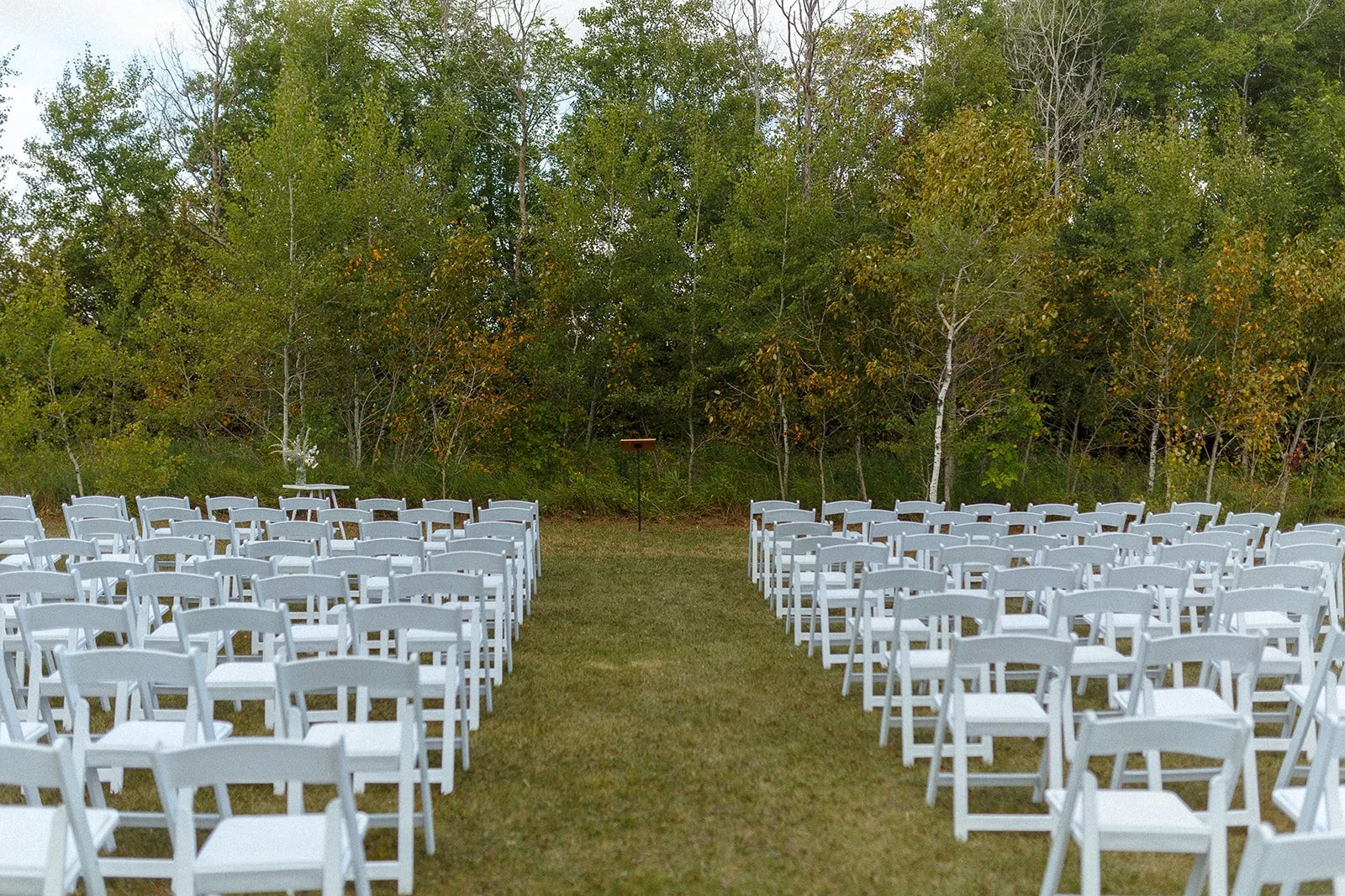 Outdoor wedding ceremony setup with white chairs arranged in rows on either side of a grassy aisle, with a wooded background and a small table at the end of the aisle.