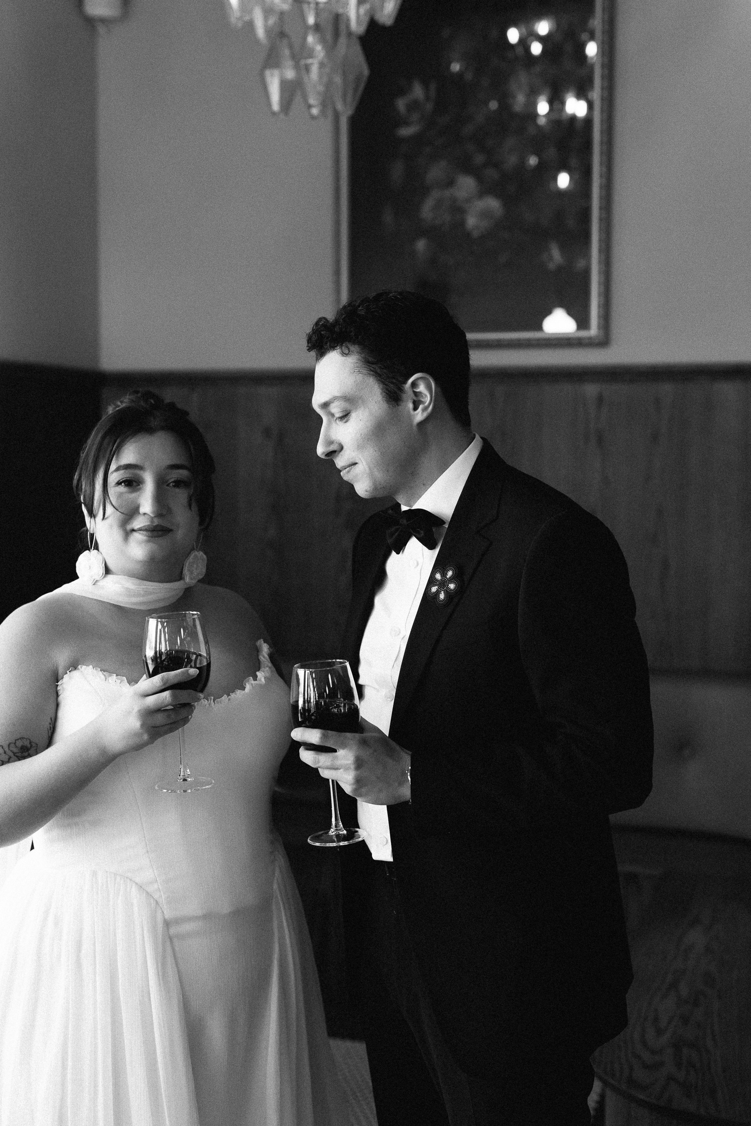 A black-and-white photo of a bride and groom holding glasses of red wine, standing close together in a formal setting, smiling softly.