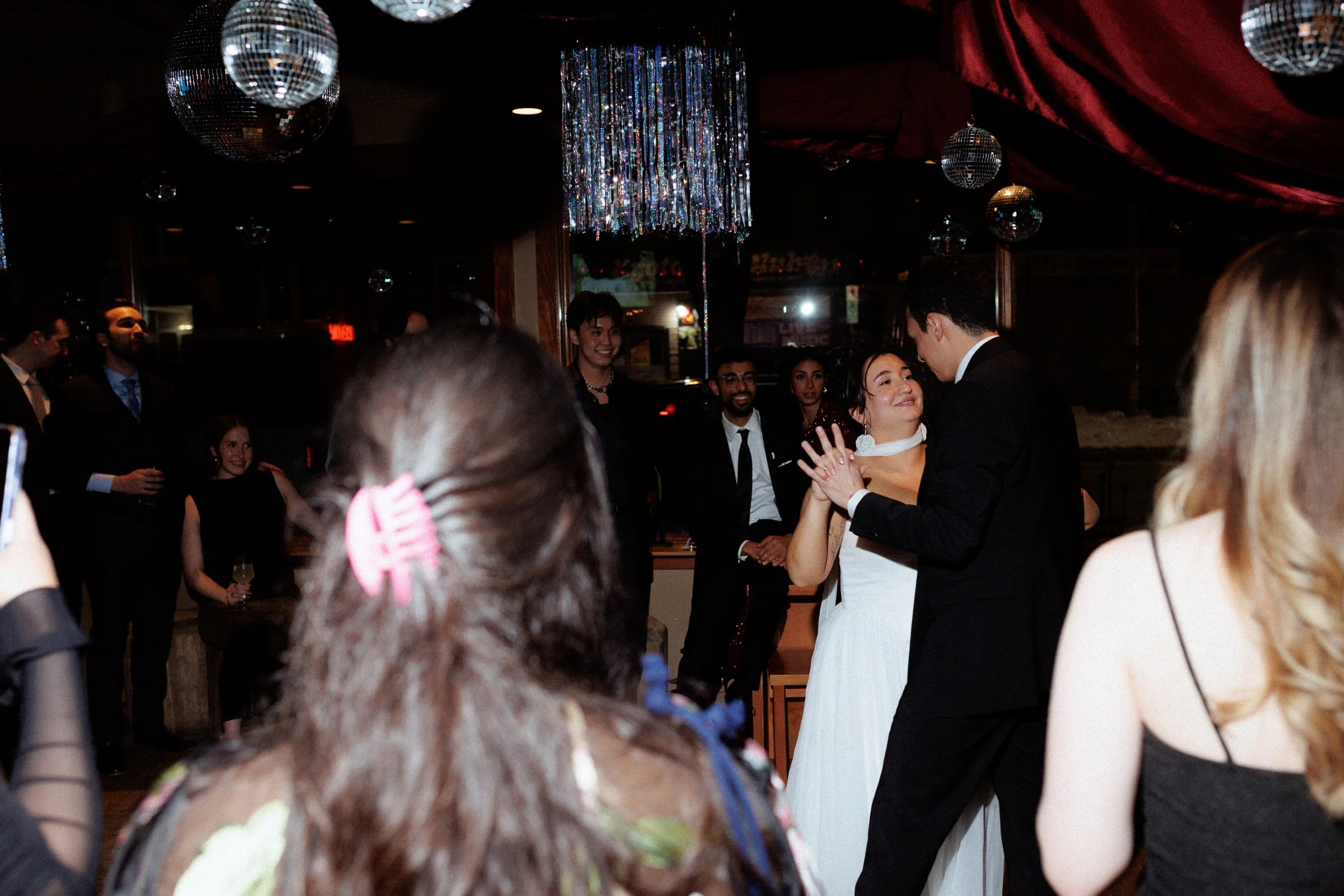 A couple dancing at a wedding reception with guests watching and celebrating in the background. Disco balls and decorations hanging from the ceiling.