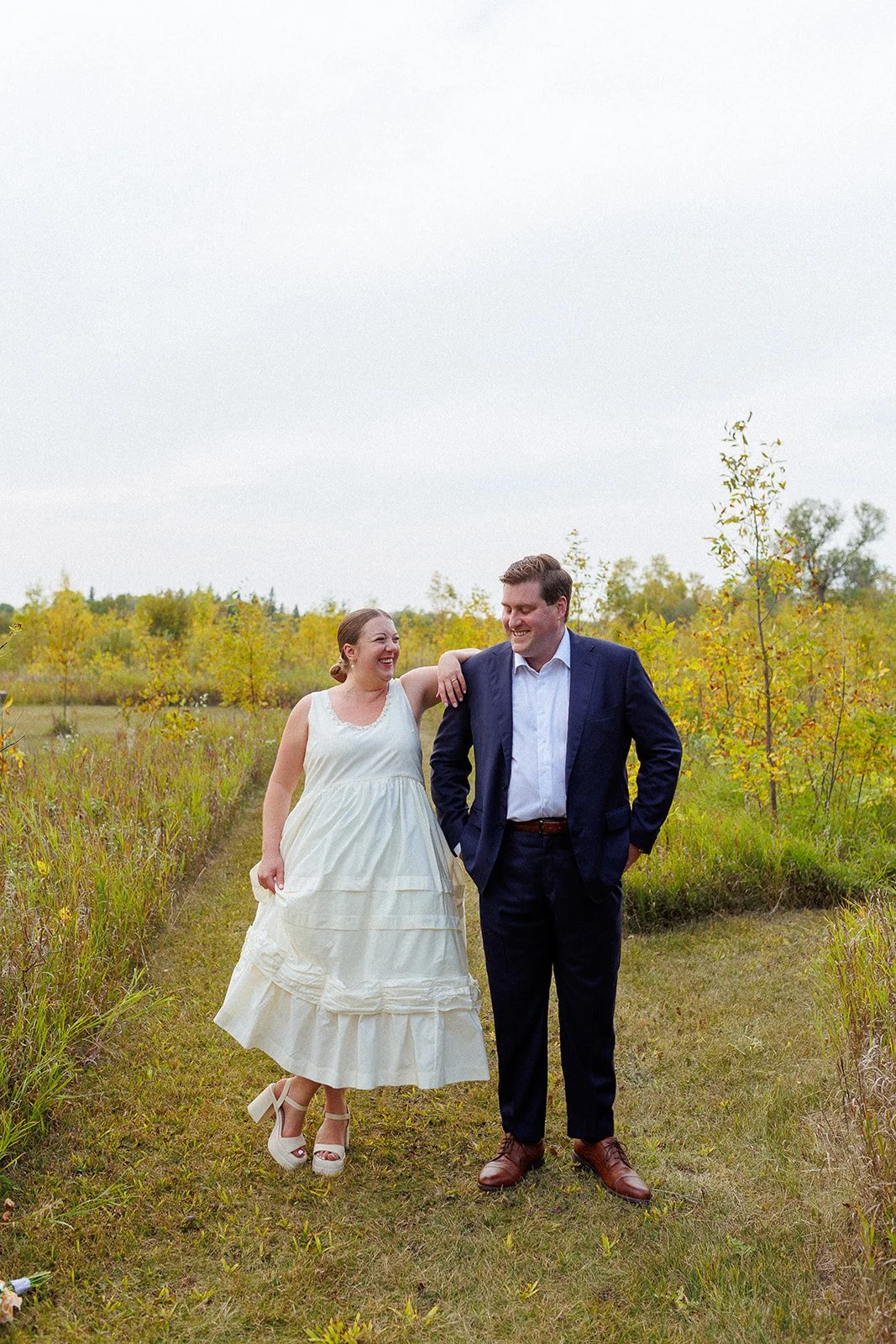 A couple is walking outdoors on a grassy path surrounded by small trees with autumn foliage. The woman is wearing a white sleeveless dress and white high-heeled sandals, and the man is dressed in a navy suit with brown shoes. They are smiling and loo