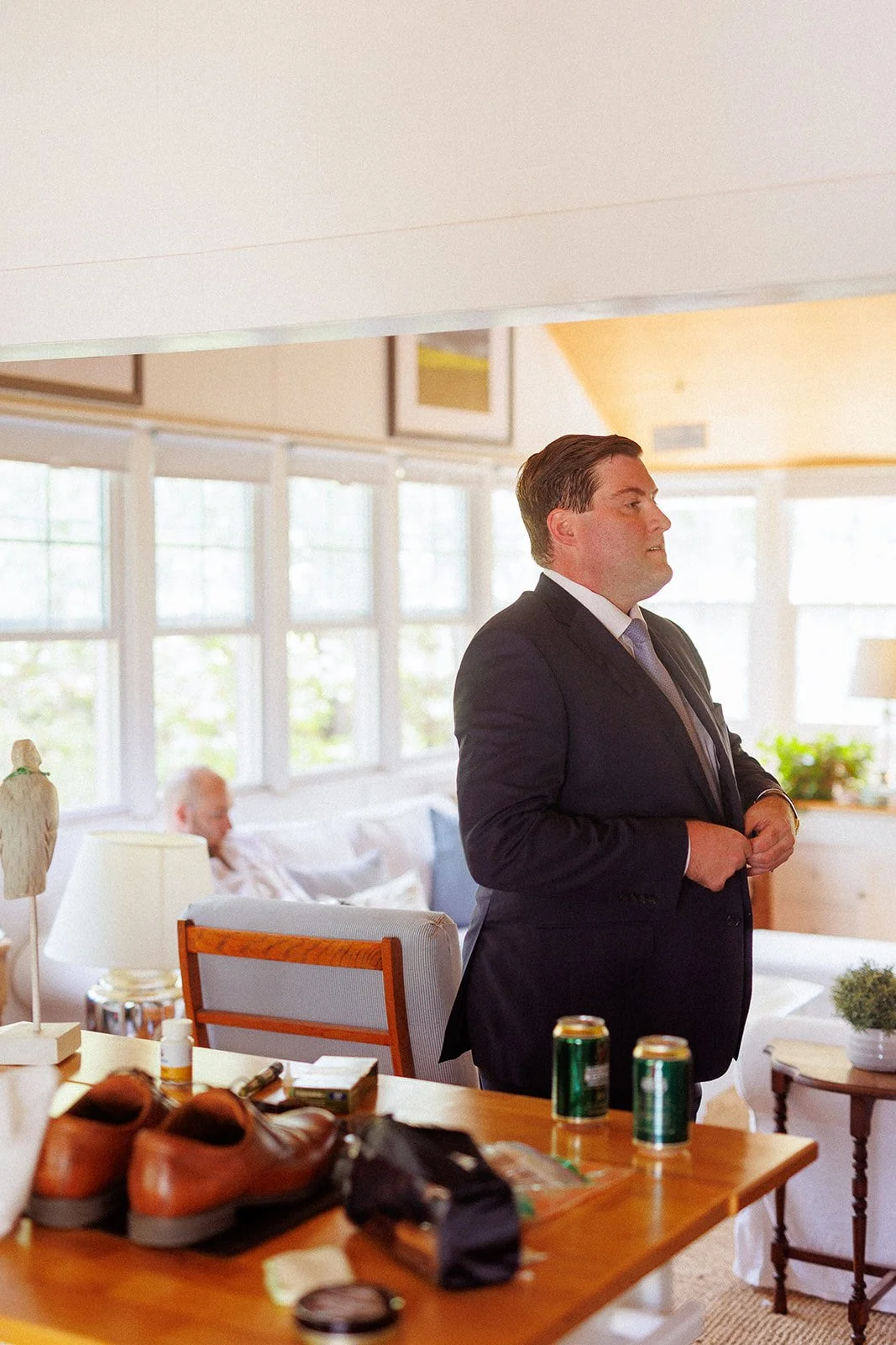 A man in a dark suit stands in a bright living room, preparing for a formal event, with an elderly woman relaxing on a sofa in the background.