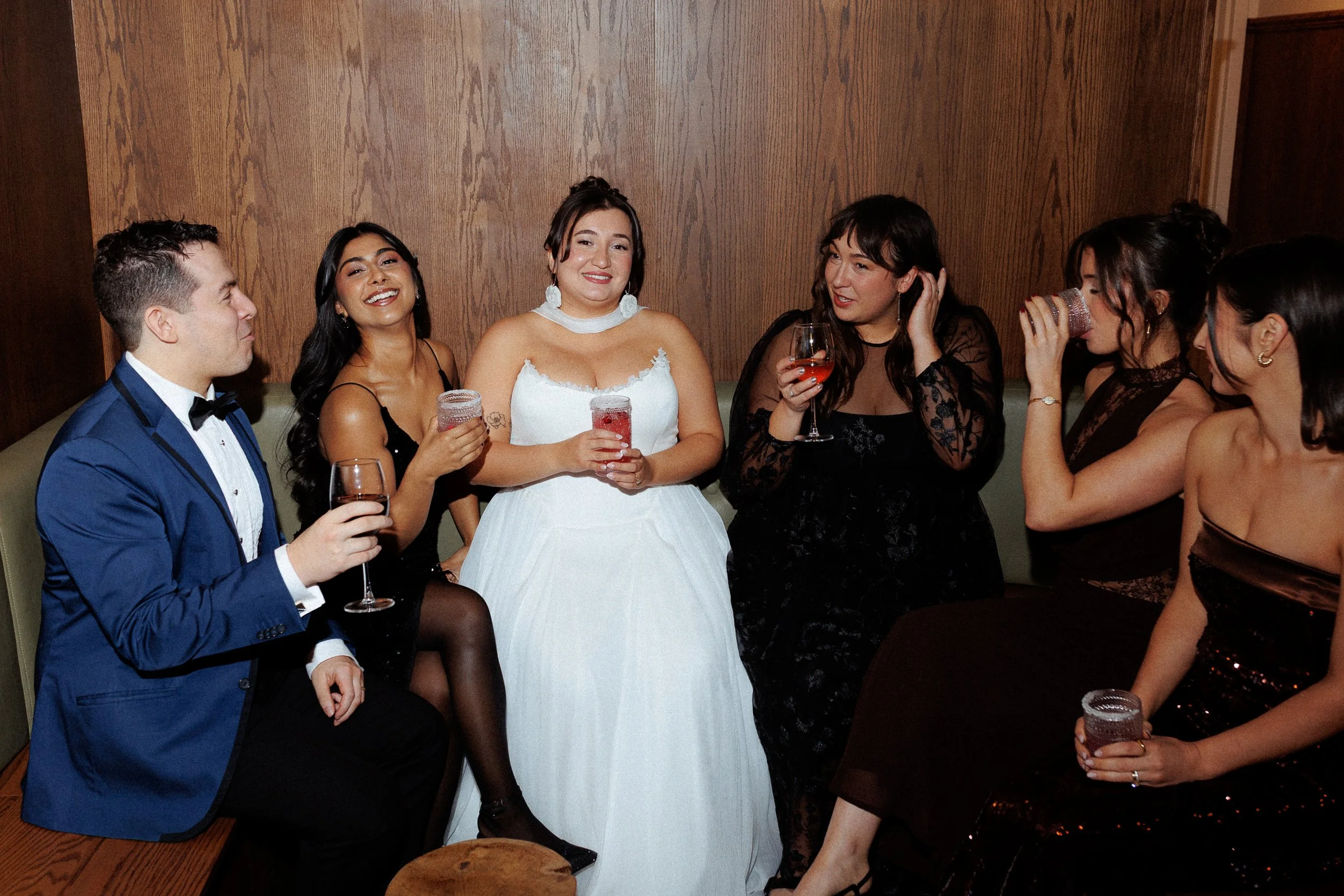 Group of six people in formal attire celebrating with drinks in a wooden-paneled room, with women wearing dresses and one man in a blue suit.