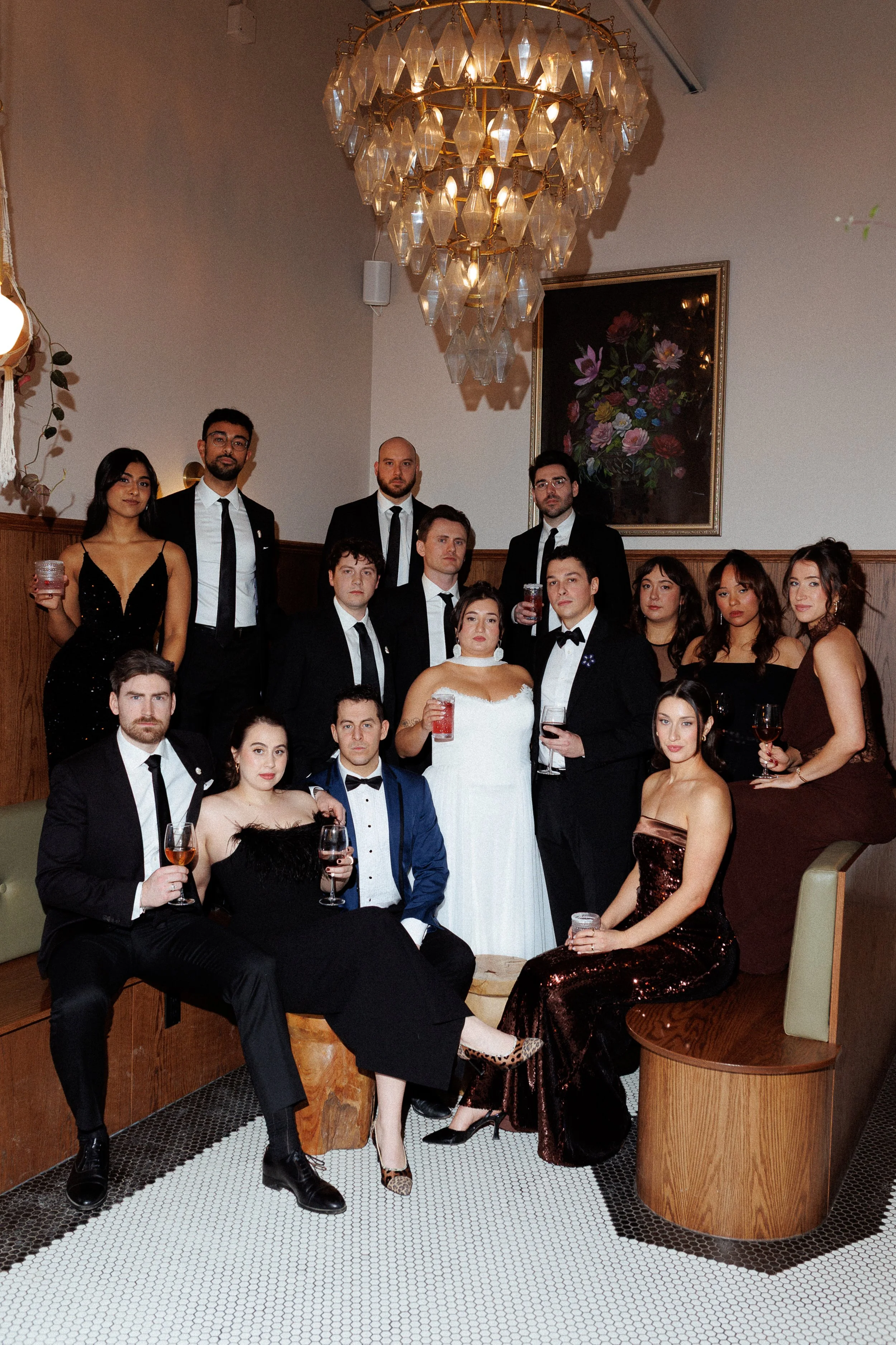 A group of 15 people dressed in formal attire, posing together at an indoor social gathering or party. They are standing and sitting around a wooden bench, with some holding drinks. The setting features a large chandelier and a framed floral painting