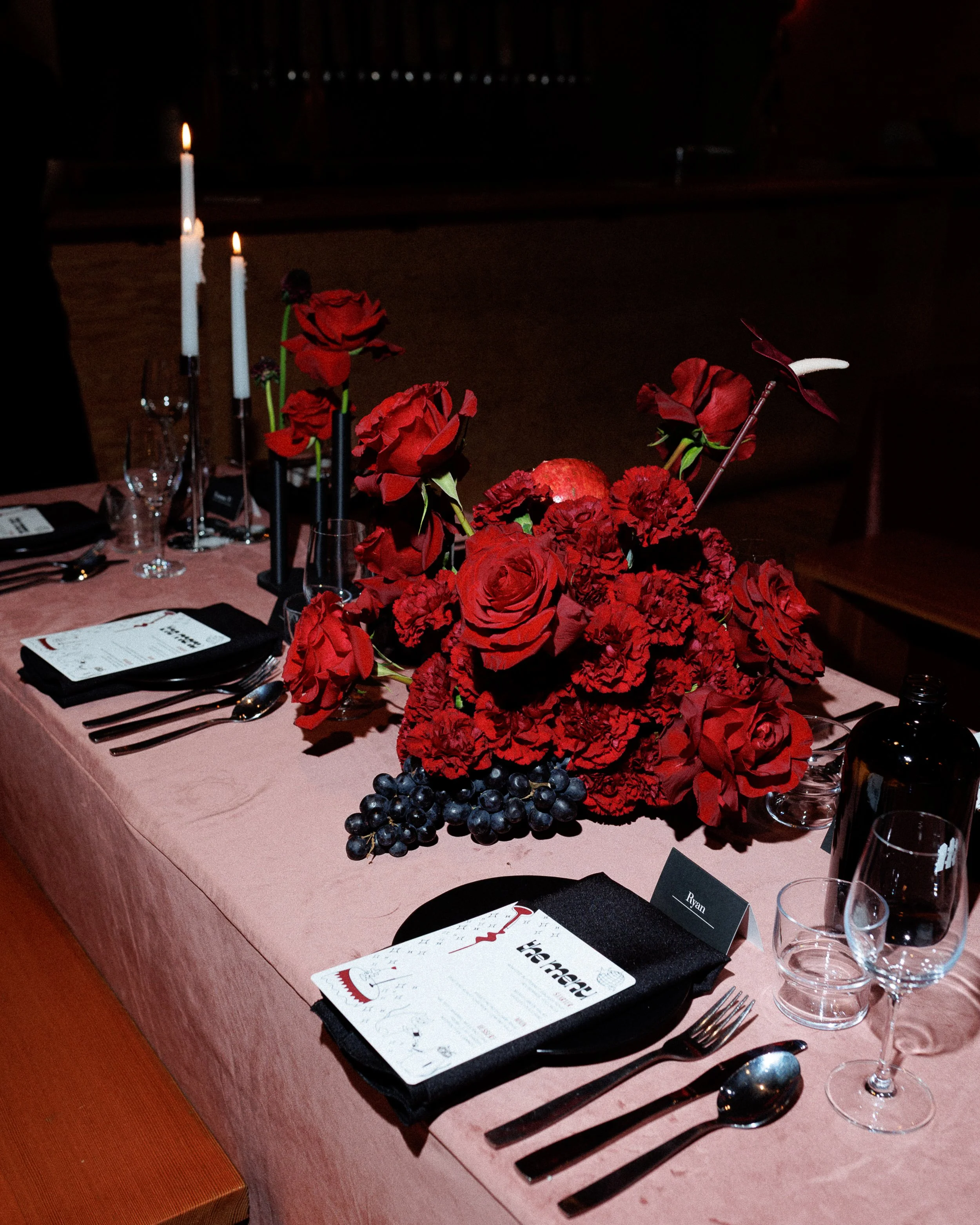 A dining table decorated with a large bouquet of red flowers, black grapes, candles, and place settings with menus, glasses, and cutlery.