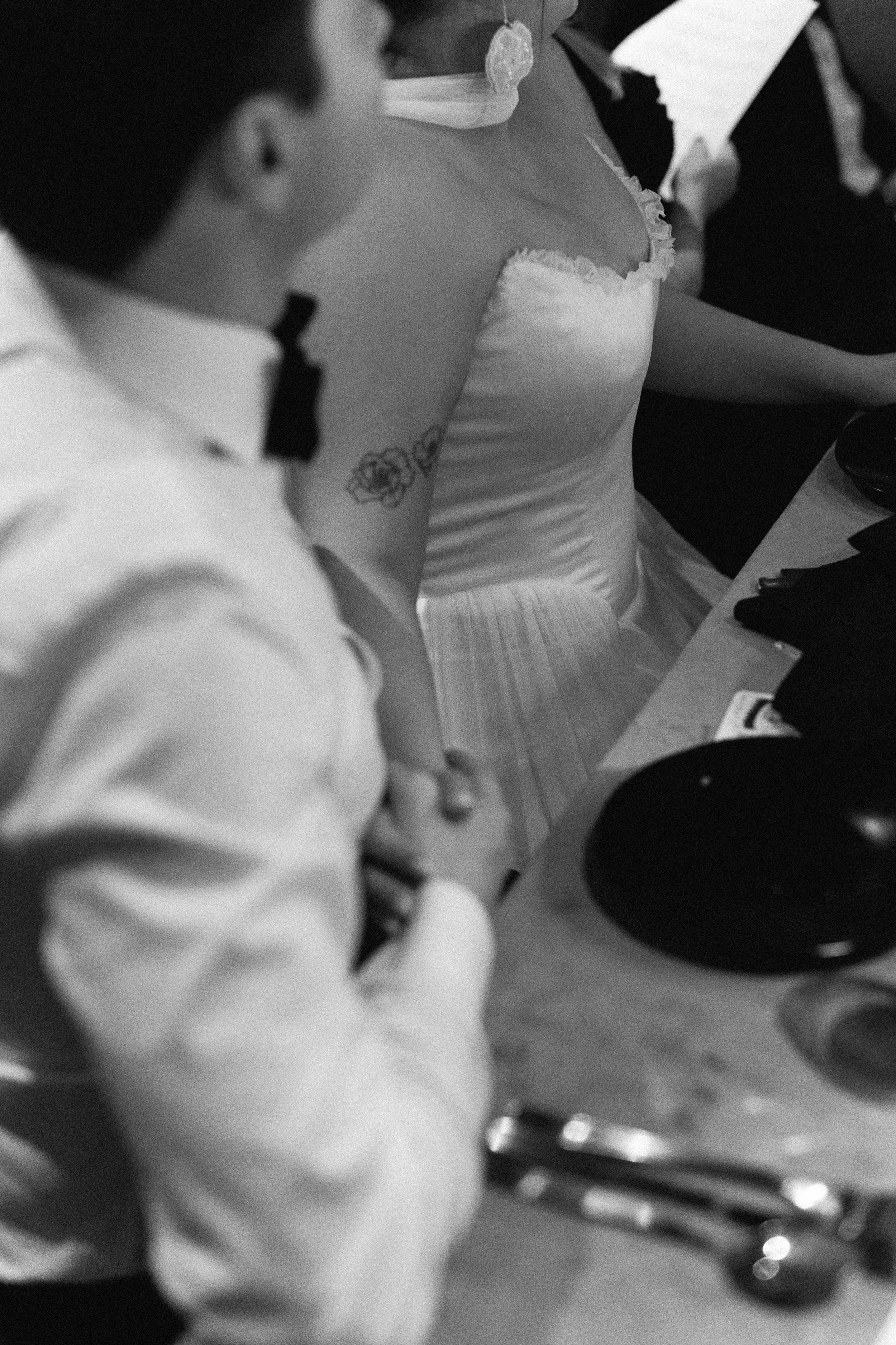 Black and white photo of people dressed in formal attire, possibly at a wedding or formal event. The person in the center is wearing a strapless dress with ruffled trim and has a tattoo of flowers on their upper arm. To the left, a person is in a whi