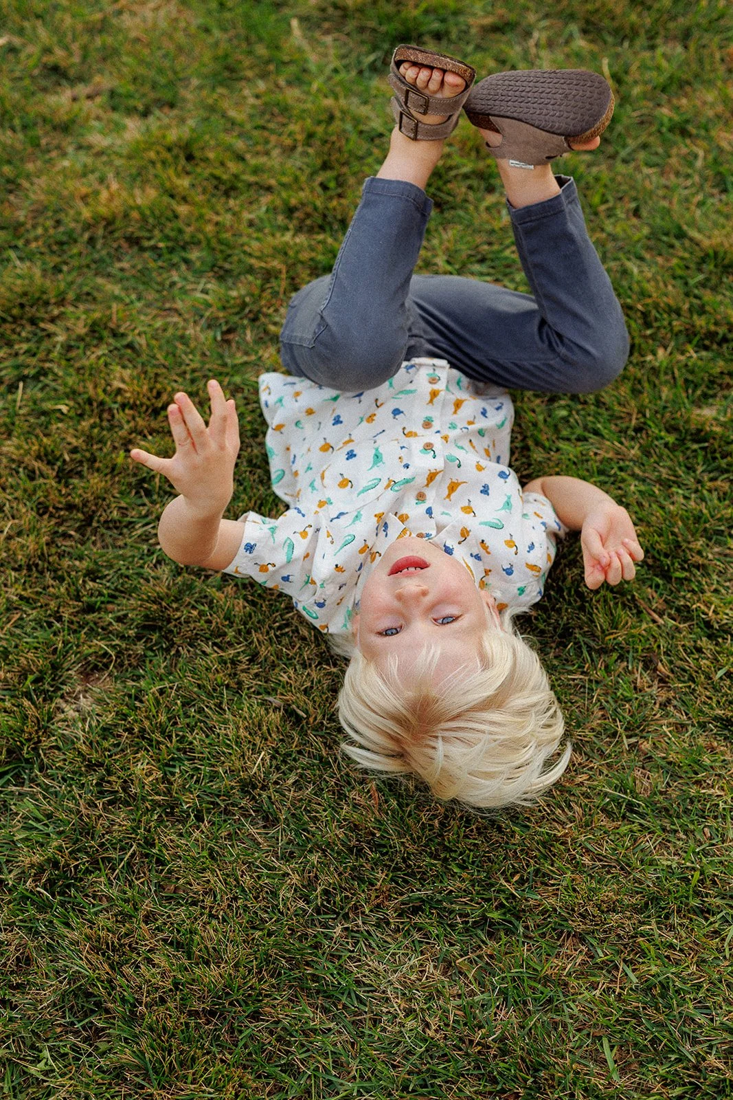 Child lying on grass with head tilted back, wearing a patterned shirt and gray pants, holding one foot up in the air.