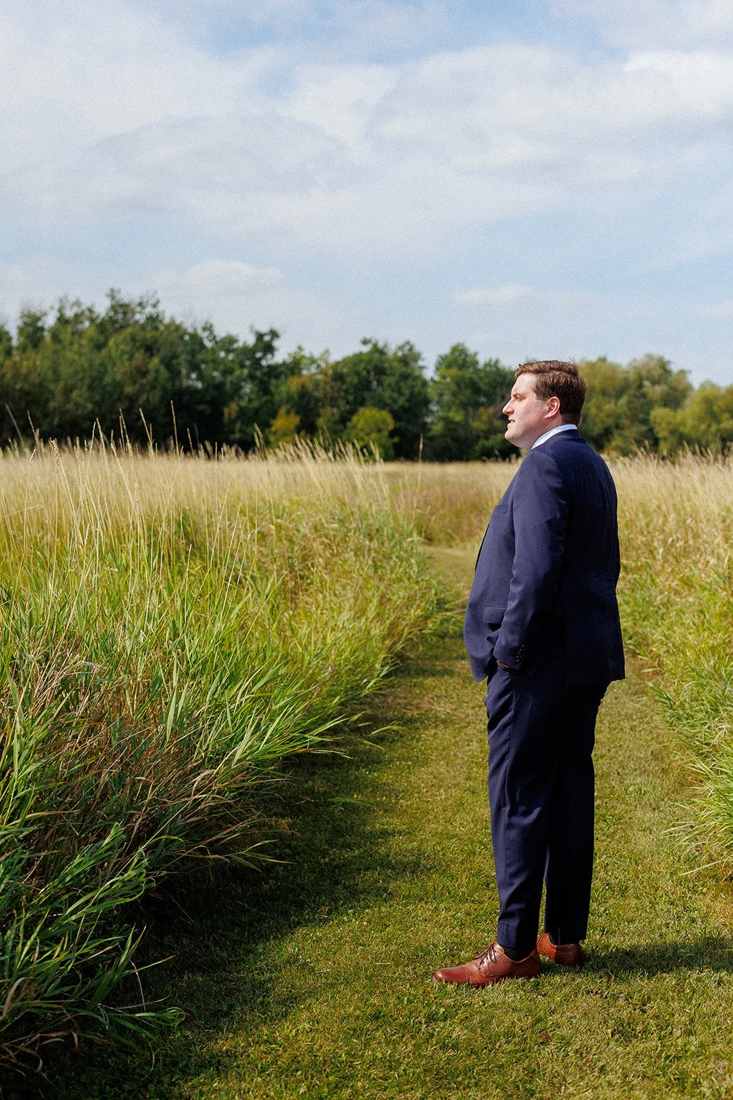 A man in a navy suit and brown shoes stands in a grassy field, looking at the distance with hands in his pockets under a partly cloudy sky.