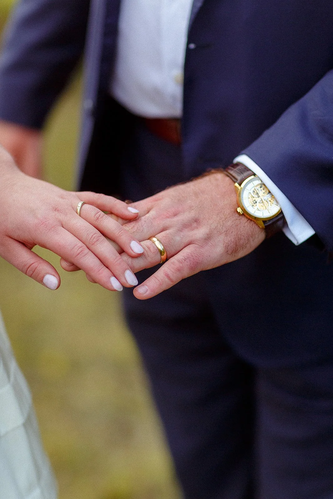 Close-up of a couple's hands intertwined, wedding rings on their fingers, with the man's hand showing a watch. The woman has short, manicured nails with white polish, and both are wearing gold rings and are dressed in formal attire.