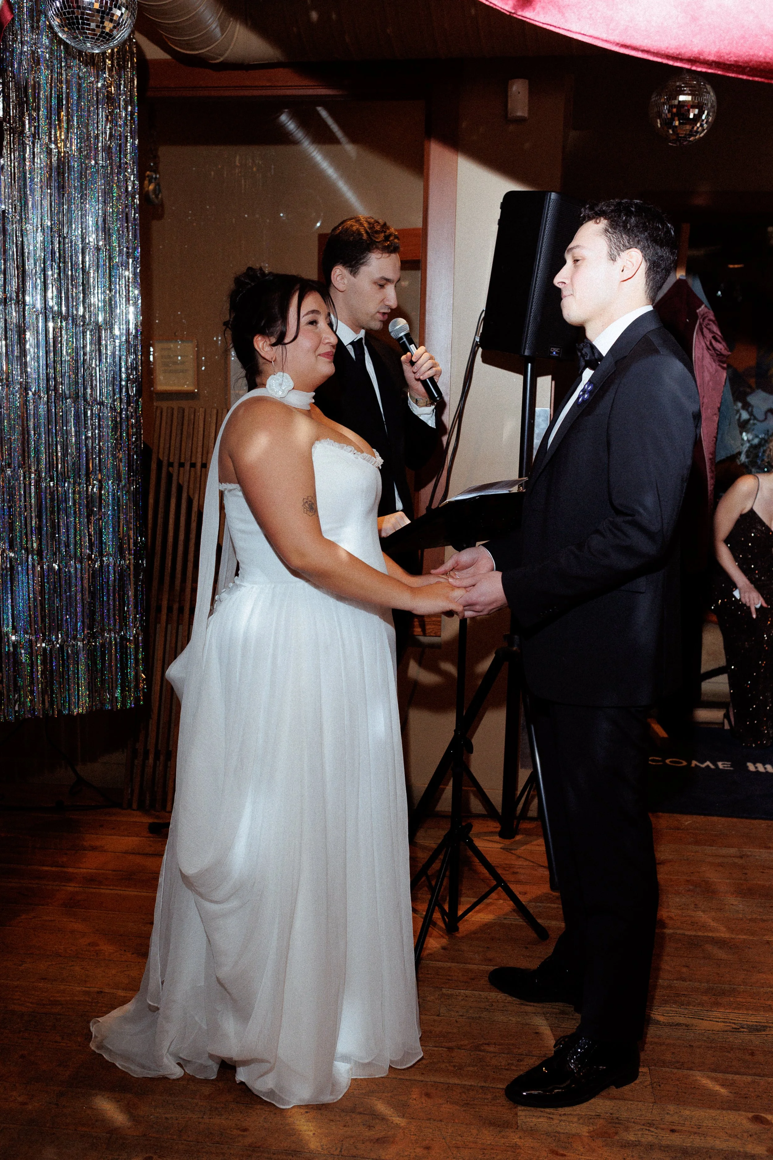 A couple getting married, holding hands and facing each other, with a woman officiant reading vows between them, in a decorated indoor venue.